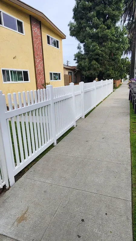 White picket fence lines a sidewalk beside a yellow apartment building with a brick accent.