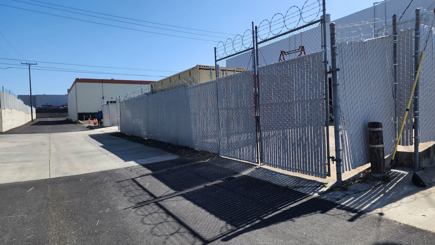 Chain link fence with barbed wire on top encloses an industrial area with buildings and an asphalt driveway.