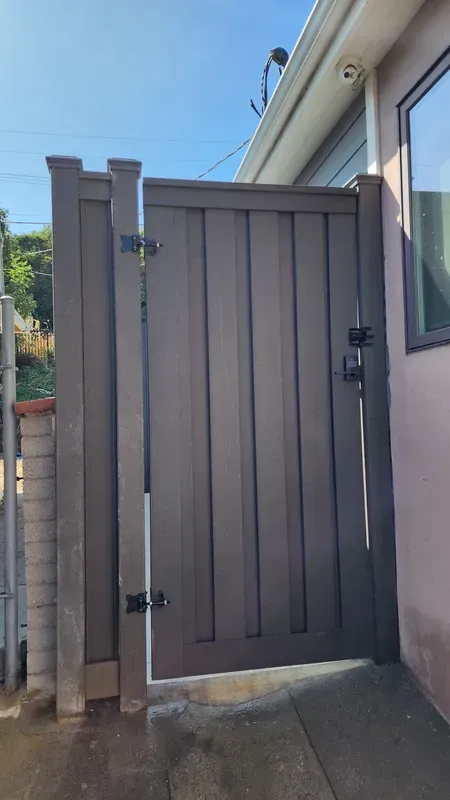 A dark brown, vertical plank gate with black hardware; next to a building with a window.