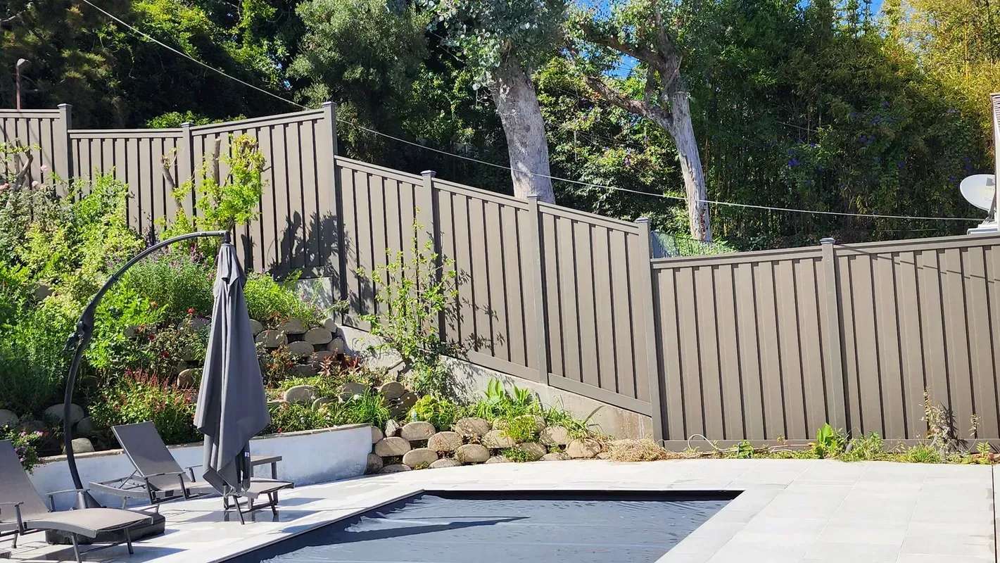 Poolside scene with fence, trees, and lounge chairs; a gray umbrella is closed.