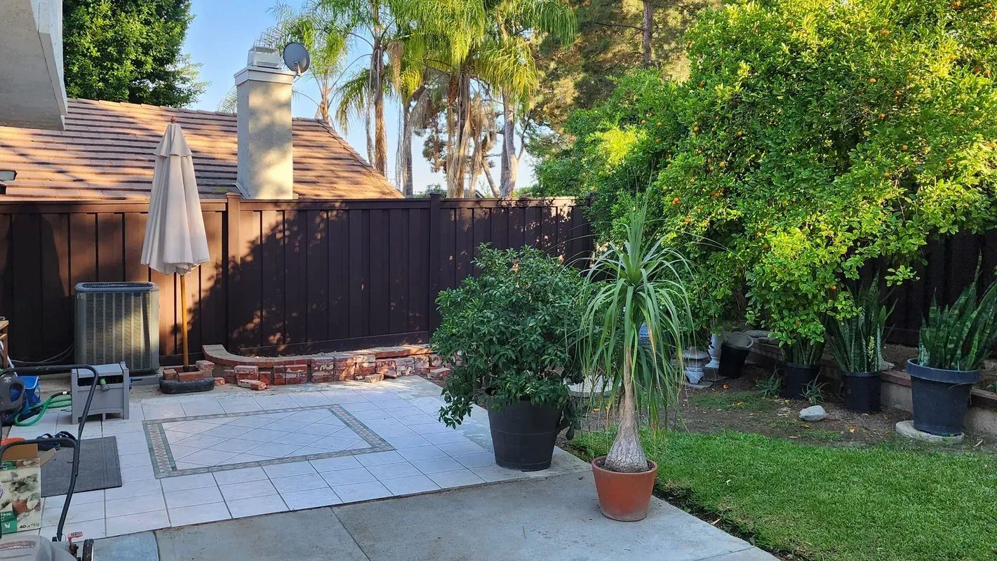 Backyard patio with brown fence, trees, potted plants, and a small patch of green grass.