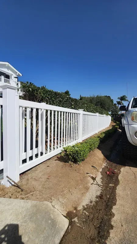 White picket fence bordering a drive, with a white car parked next to it under a clear blue sky.