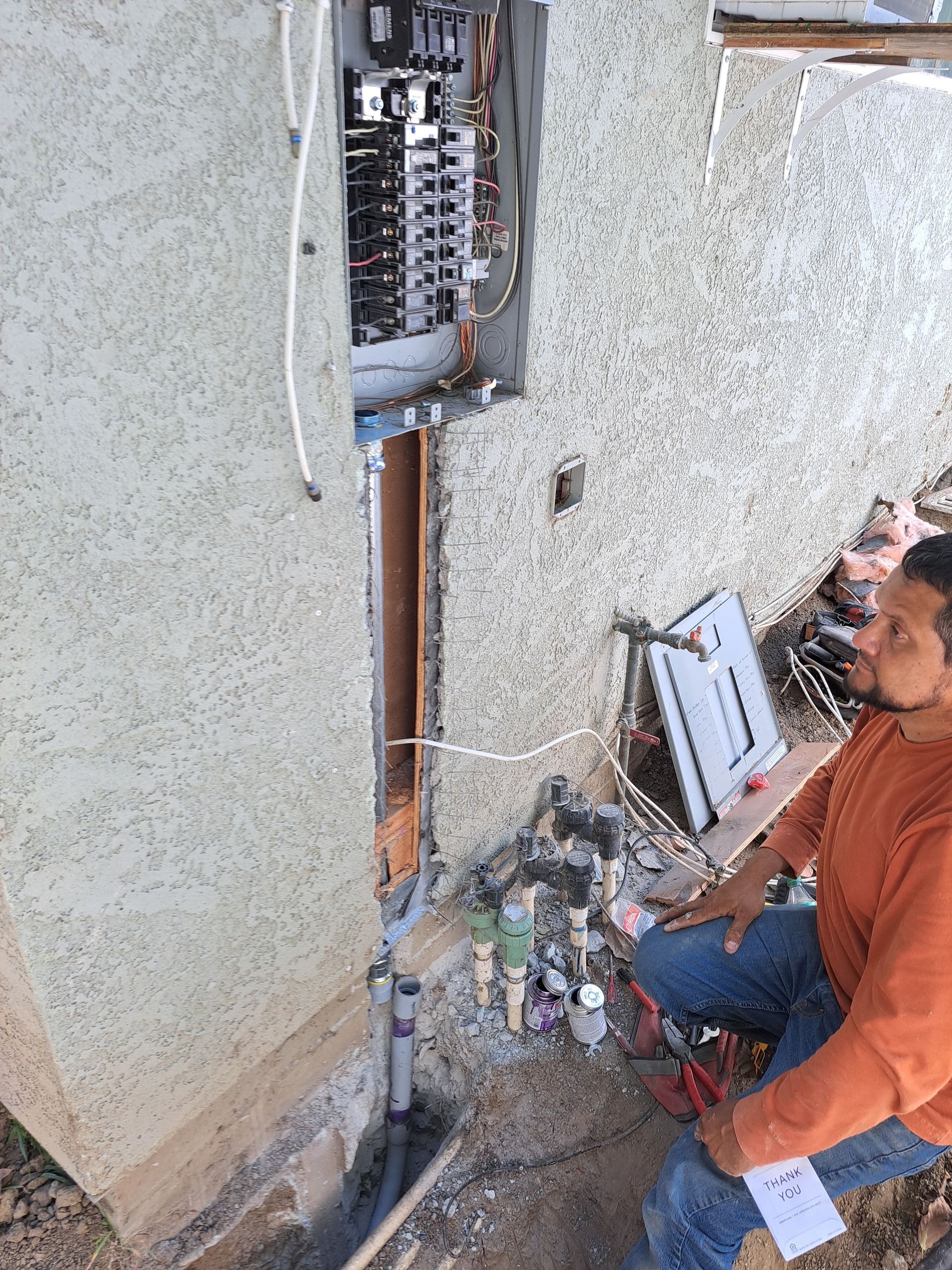 A man is sitting on the side of a building looking at an electrical box.