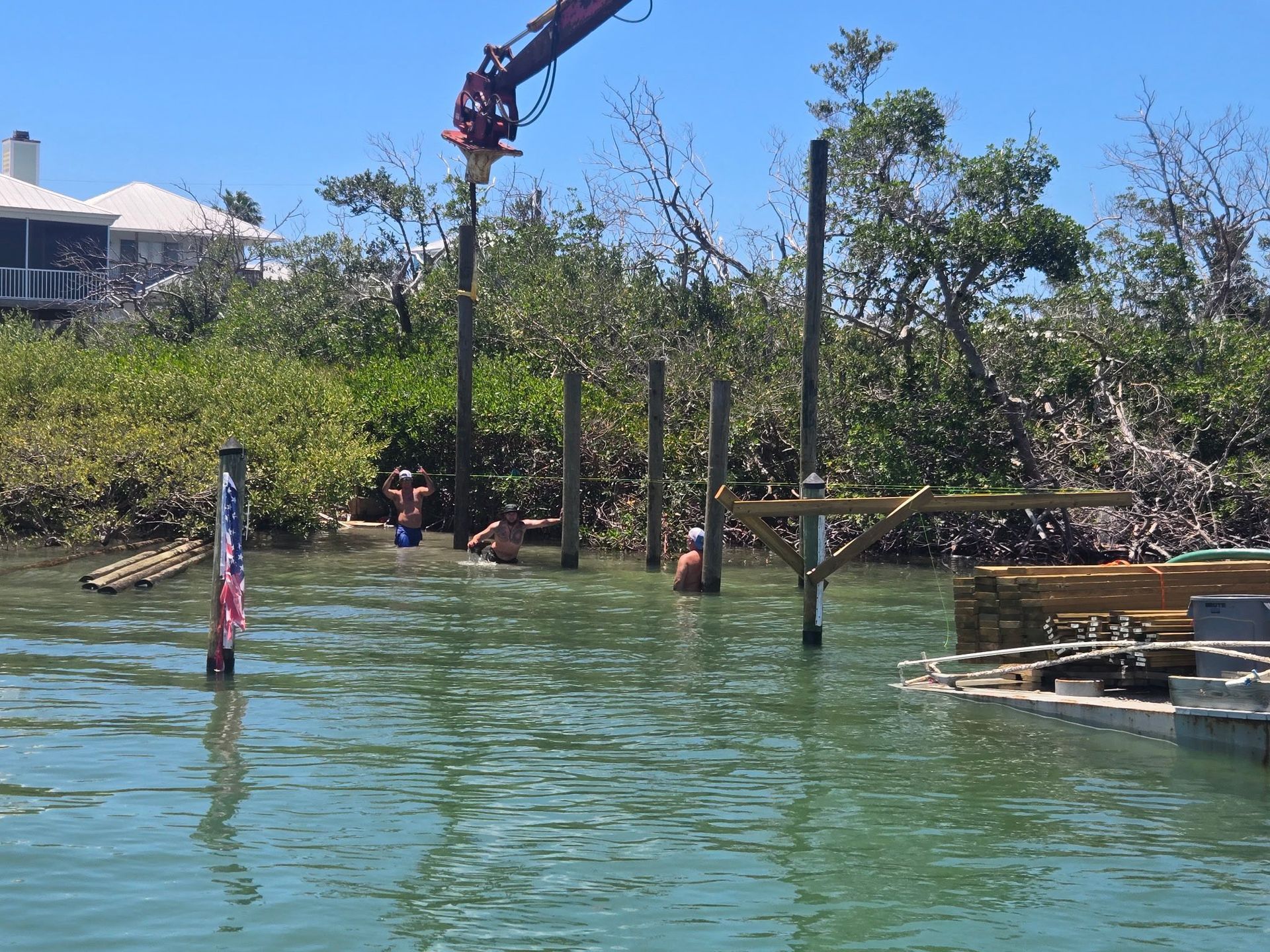People standing by a green waterfront near mangroves, with a small dock and crane in the background