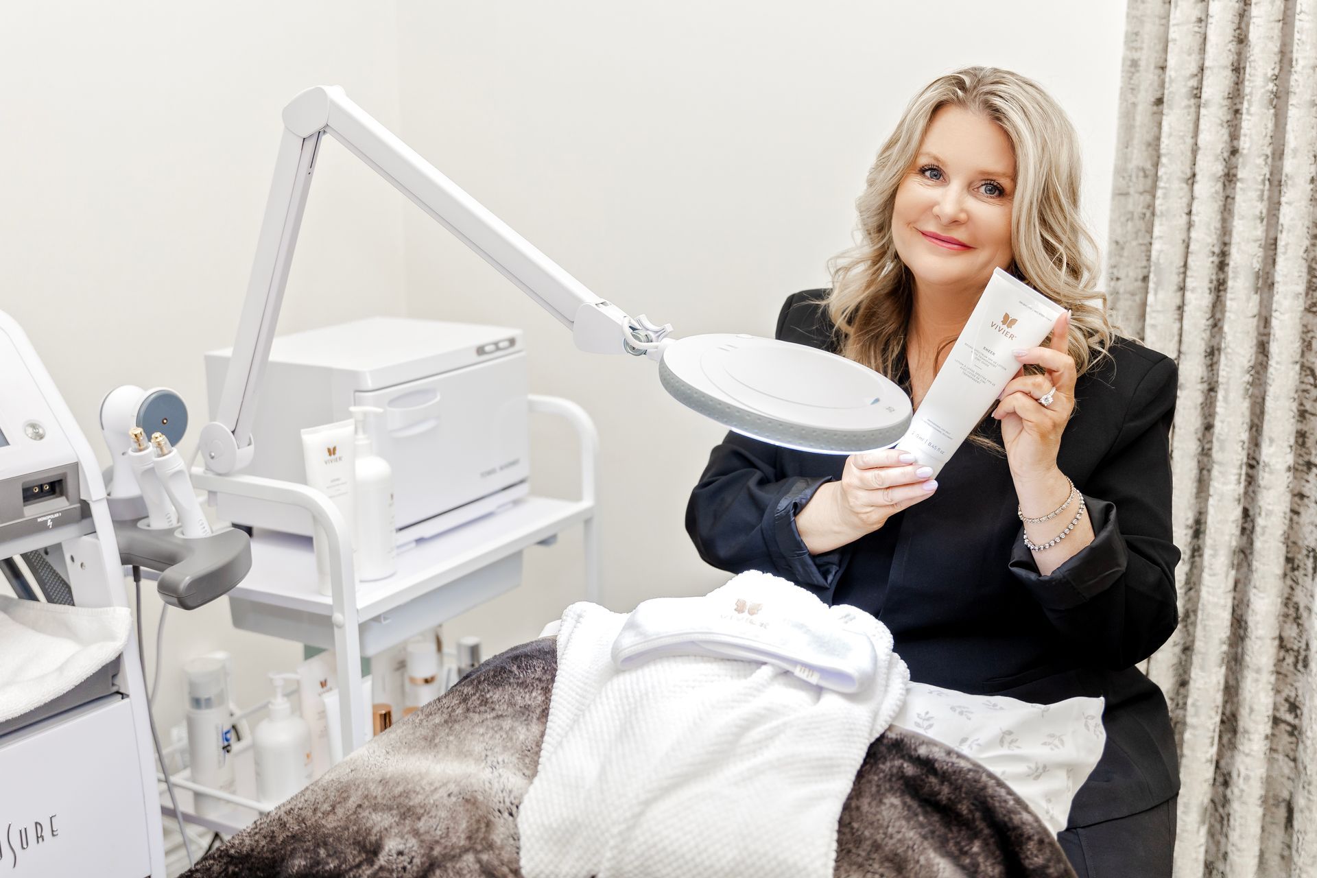 A woman is sitting on a bed holding a magnifying glass and a bottle of lotion.