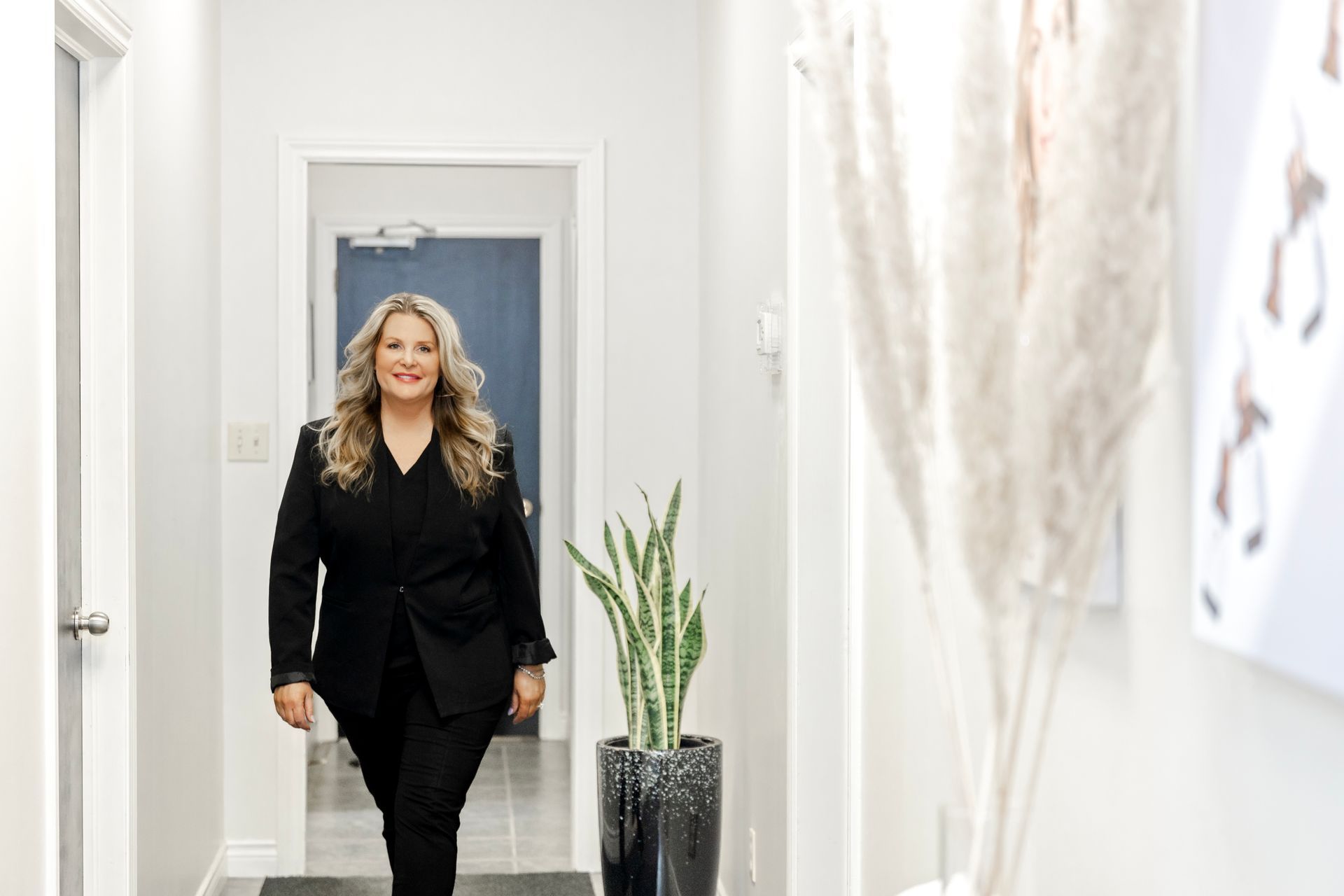 A woman in a black suit is walking down a hallway next to a potted plant.