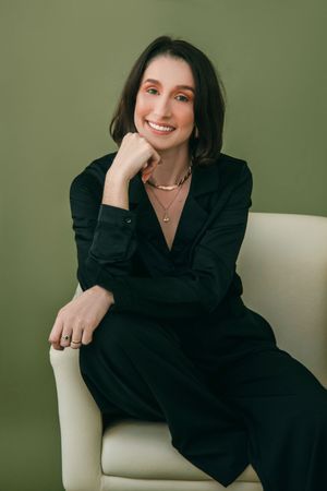 A woman in a black dress is sitting in a chair with her hand on her chin.