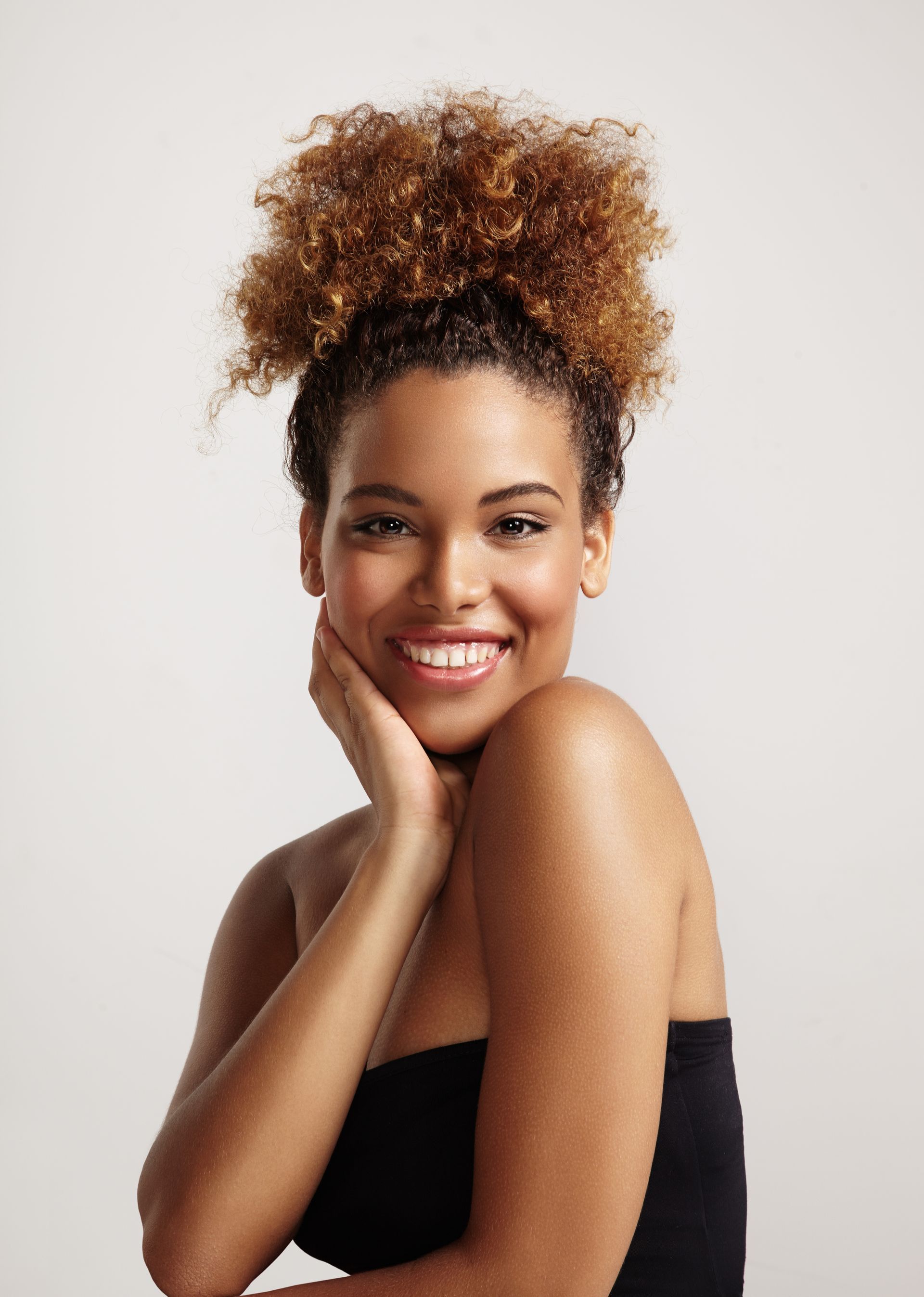 A woman with curly hair is smiling and touching her face.