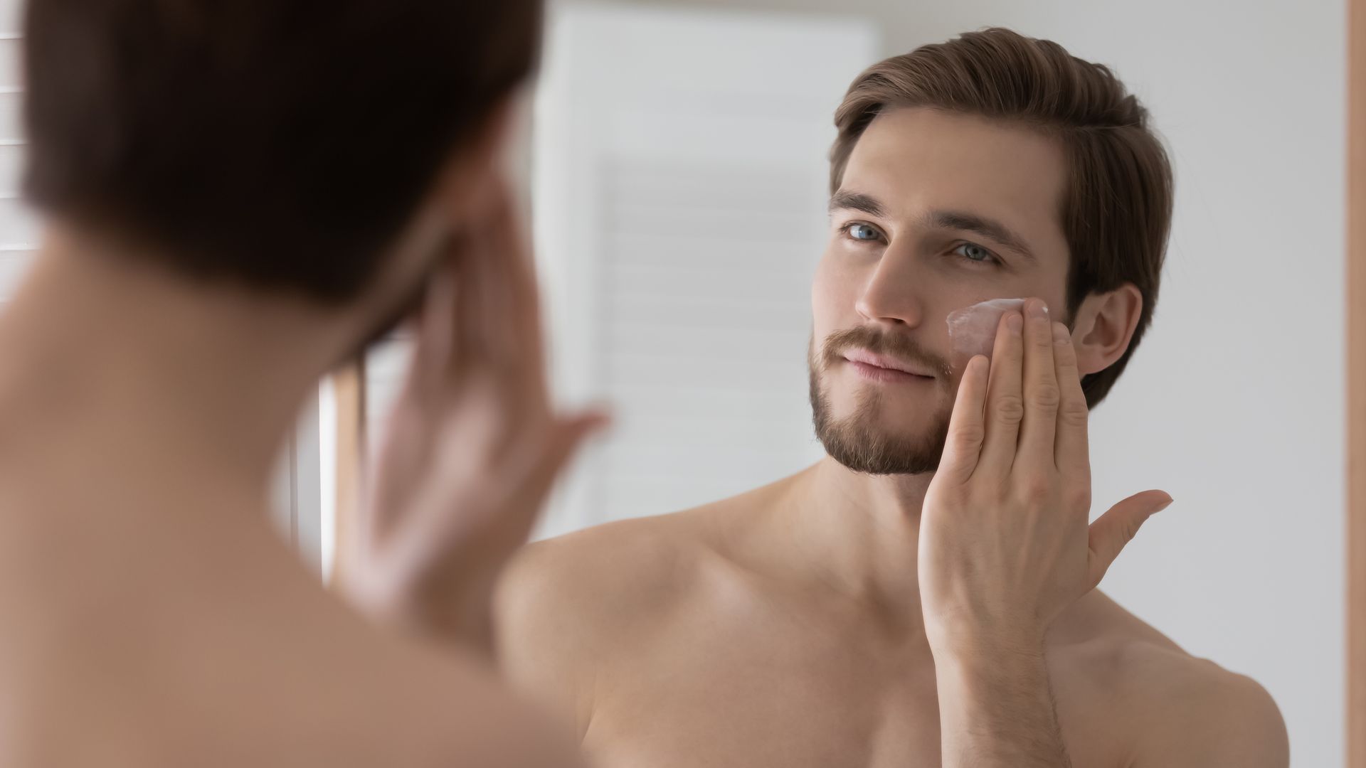 Young man applying face moisturizer while looking in the mirror, enjoying his skincare routine.
