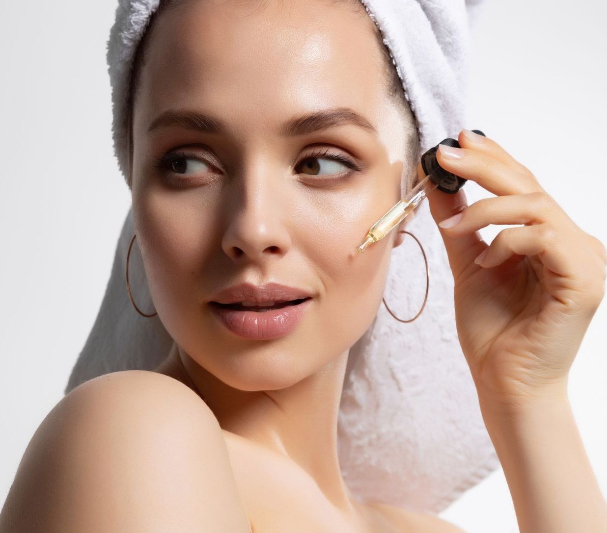 A woman with a towel wrapped around her head is applying a serum to her face.