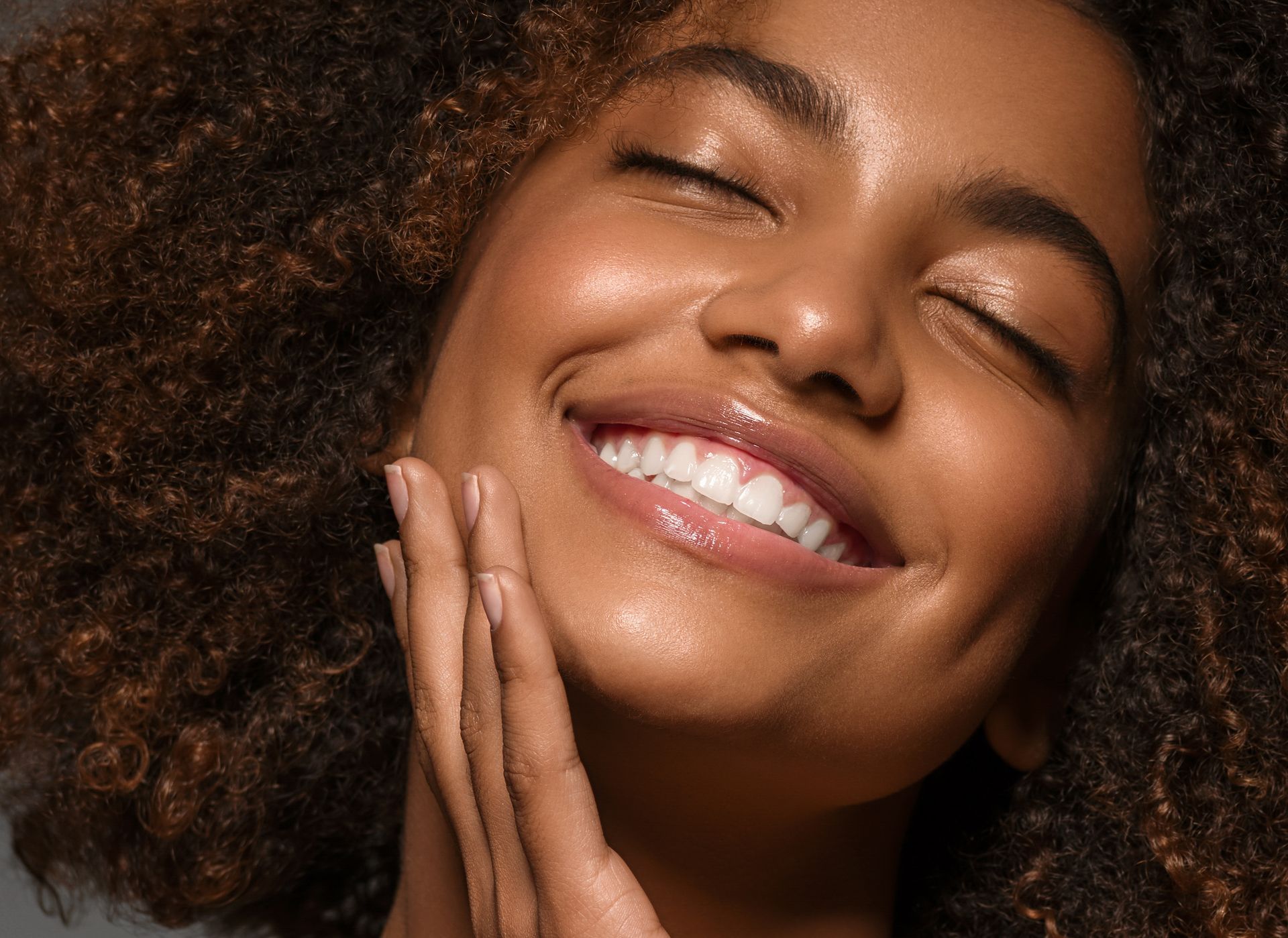 A woman with curly hair is smiling with her eyes closed and touching her face.