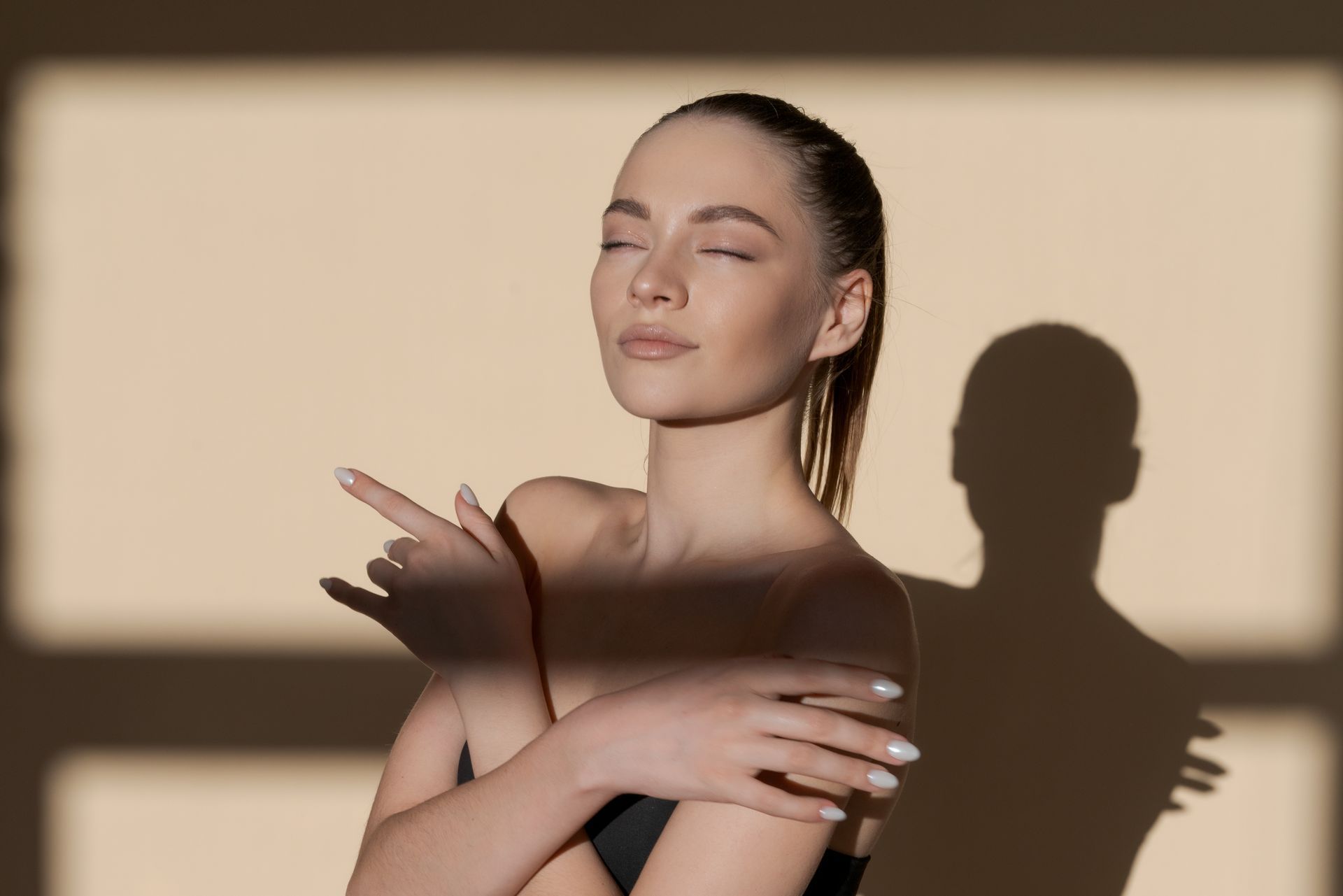 A woman is standing in front of a window with her eyes closed and her shadow on the wall.