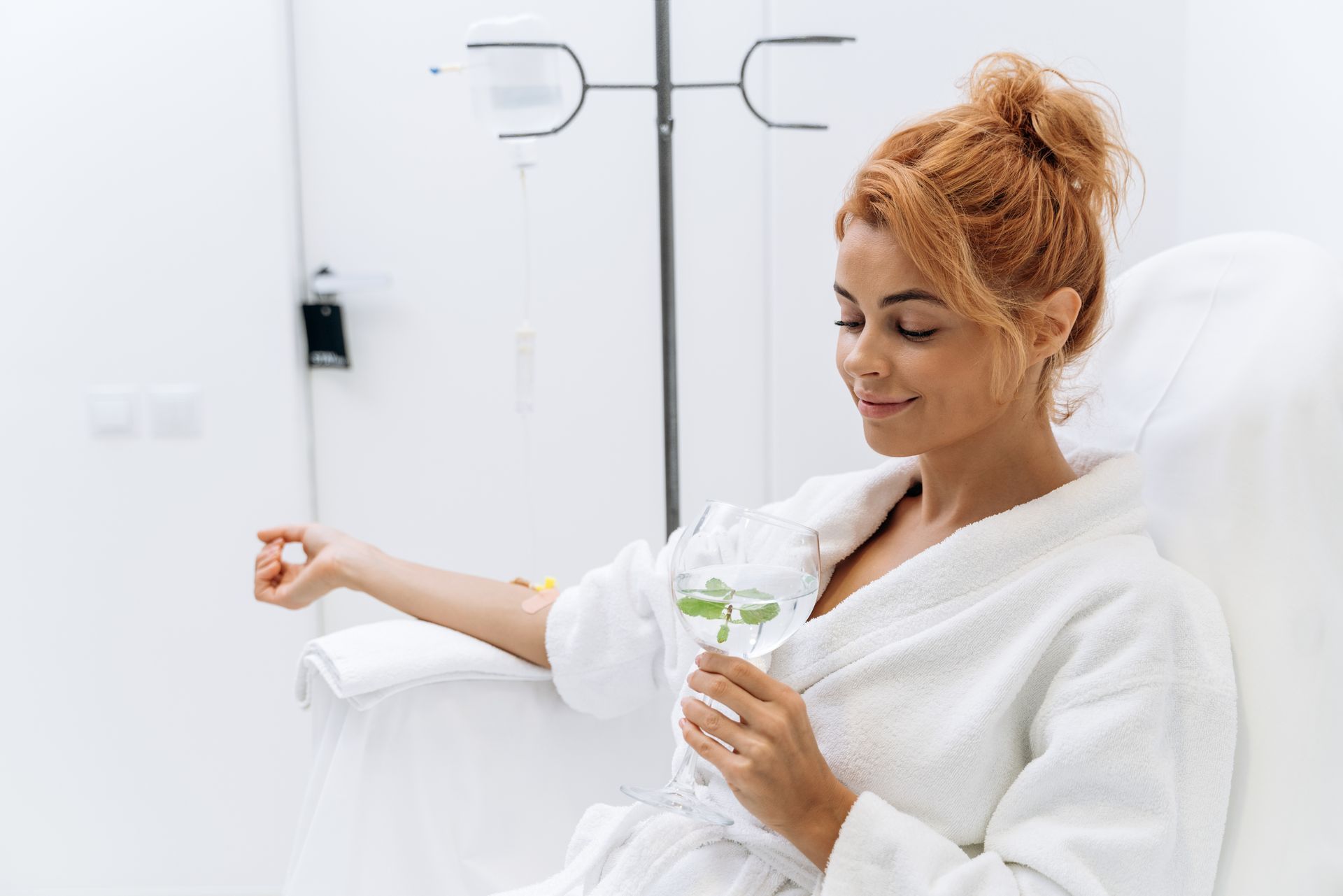 A woman in a bathrobe is sitting in a chair holding a glass of water.