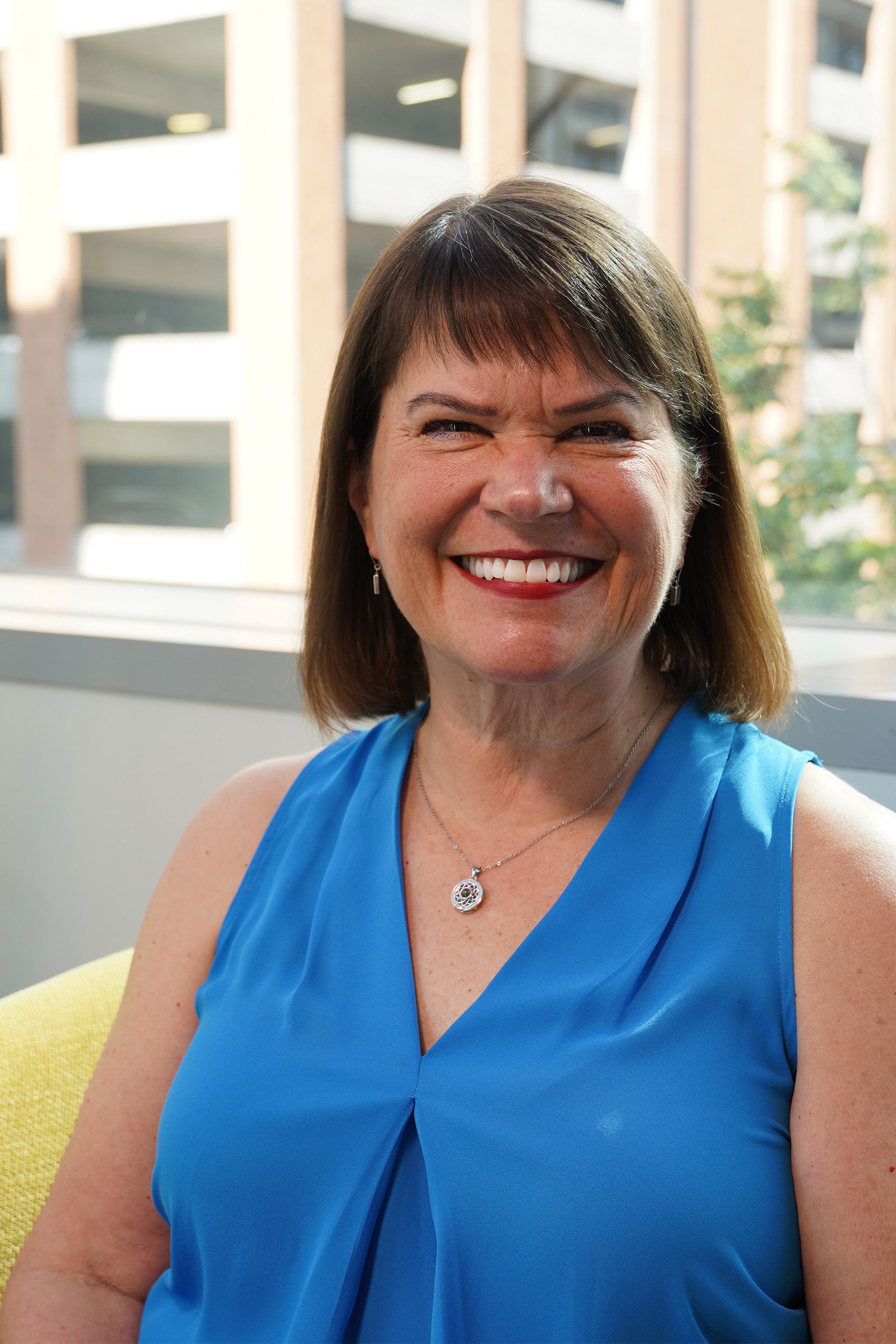 A woman in a blue shirt is smiling and sitting in front of a window.