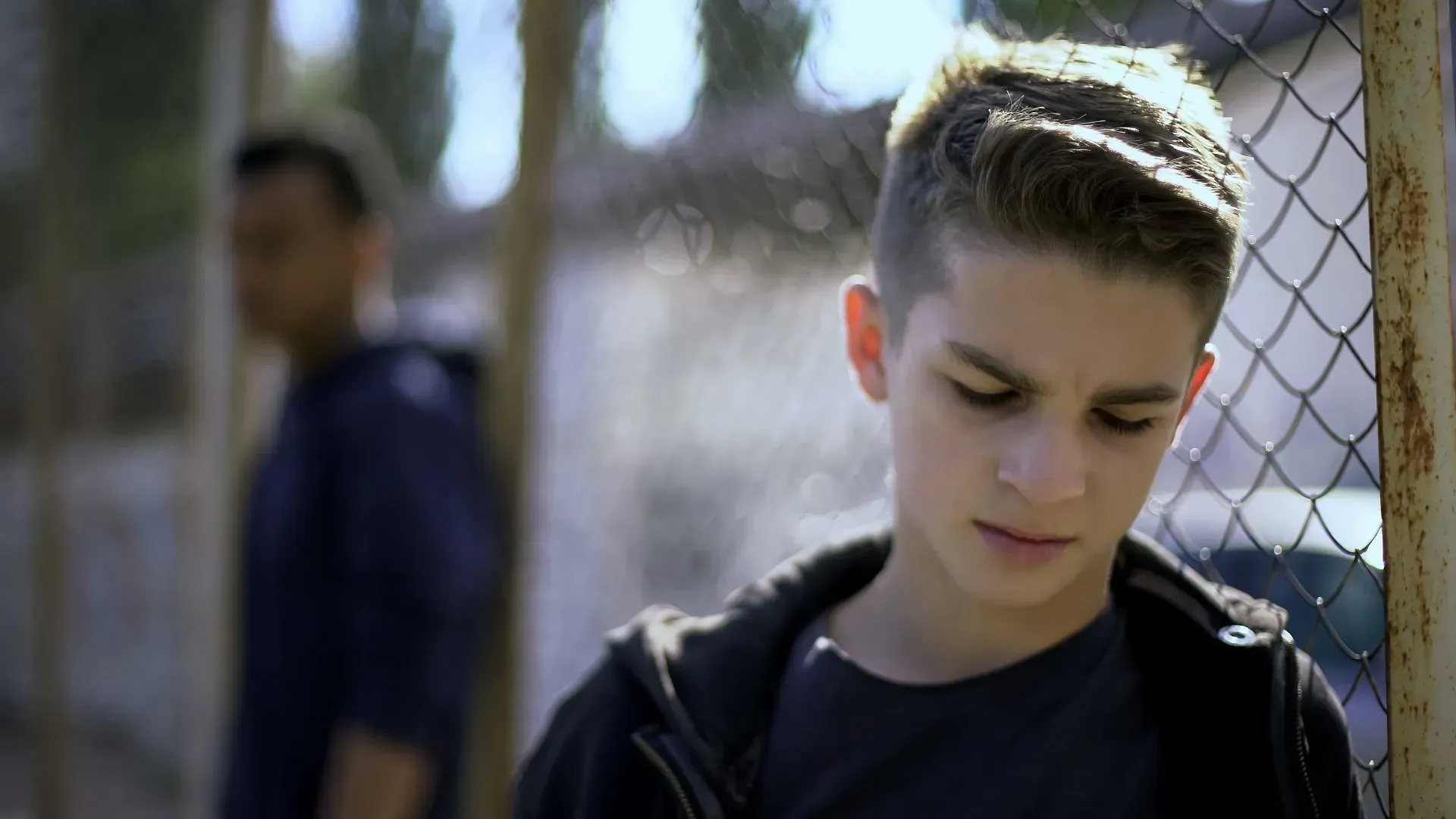 Teen boy looks down near a chain-link fence. Another figure stands blurred in the background.
