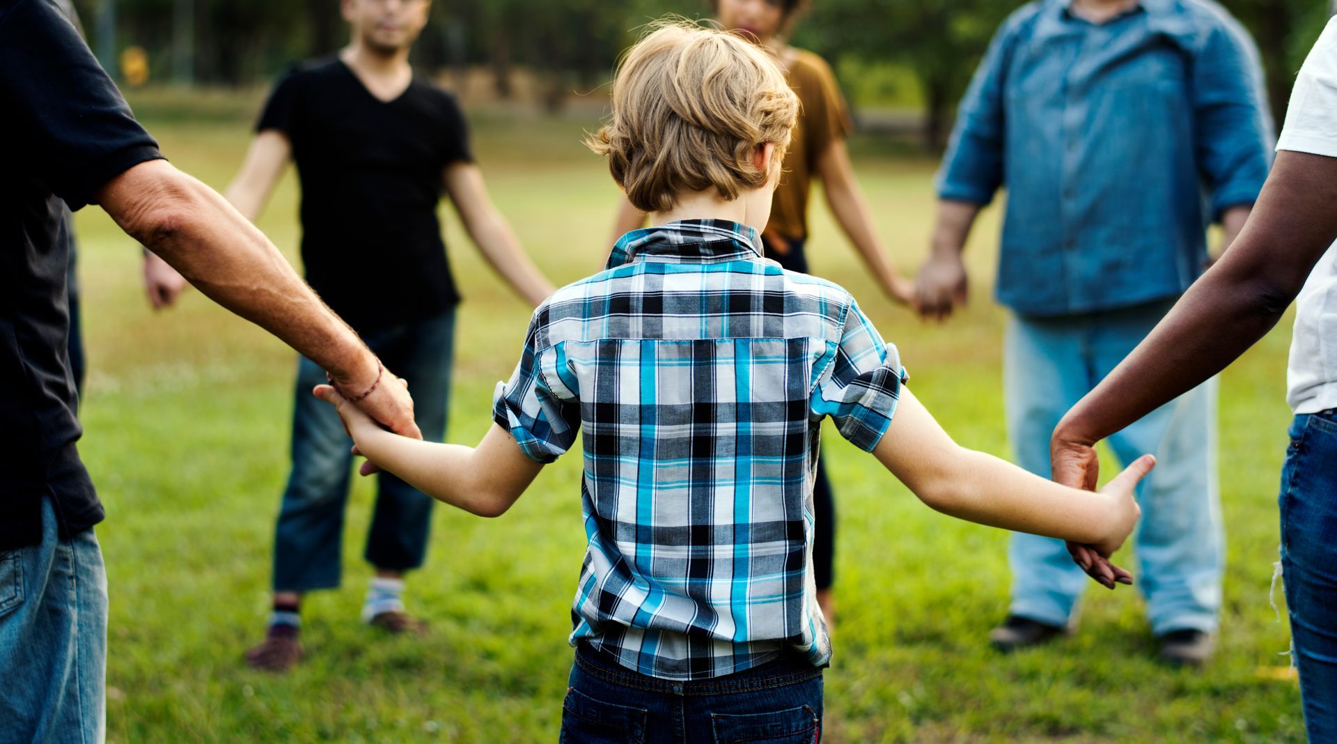 People holding hands in a circle on grass, boy in center wearing plaid shirt.