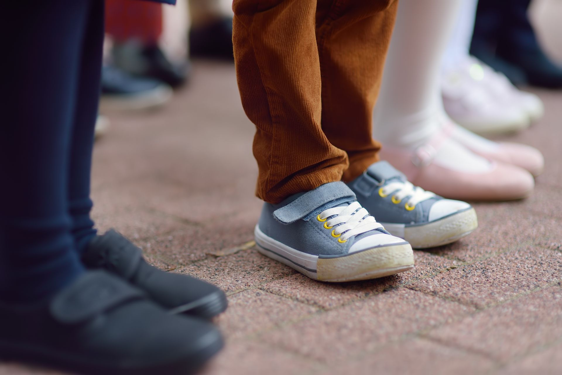 Children's legs and feet in various shoes, standing on brick paving. One pair wears blue sneakers.