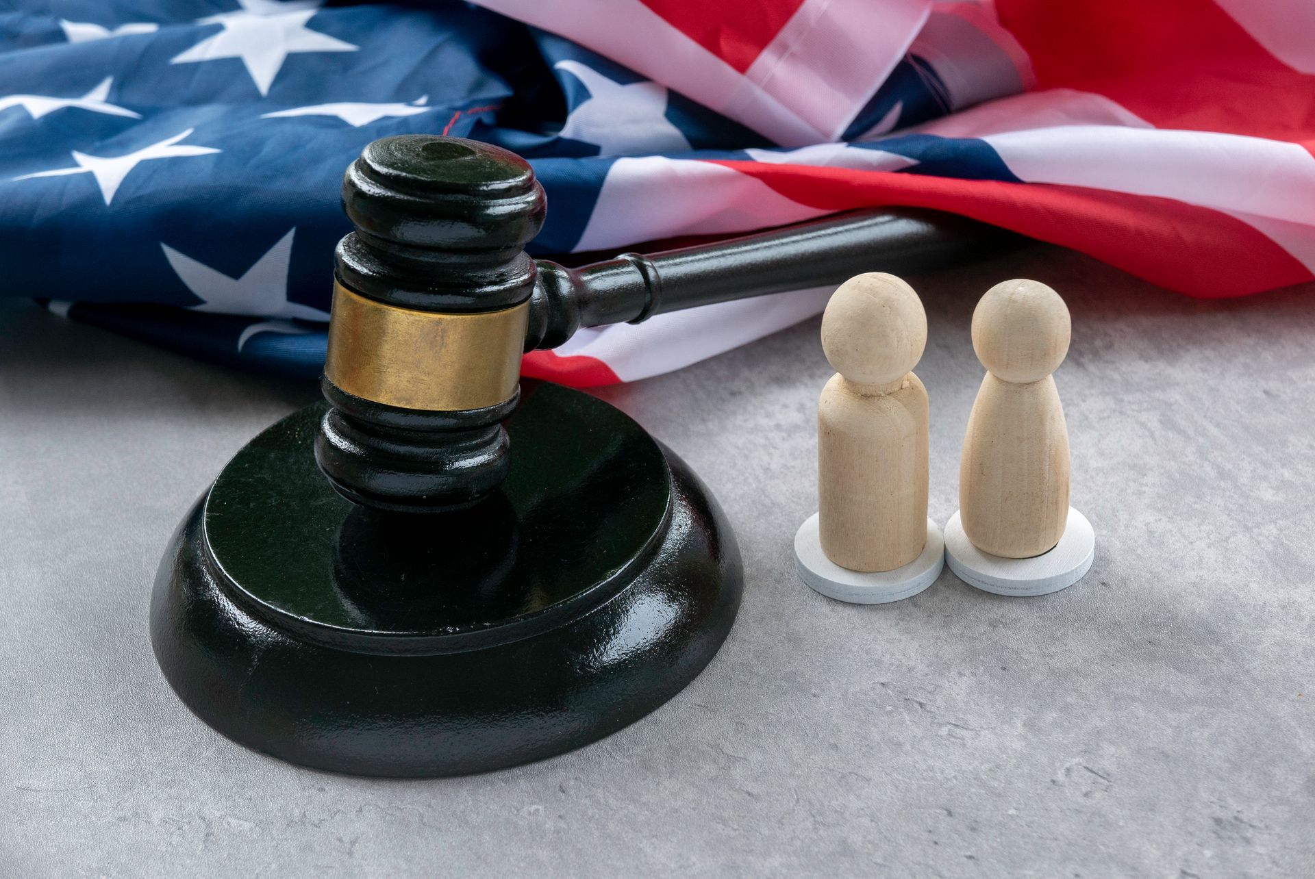 Gavel and two wooden figures on a concrete surface with the US flag in the background.