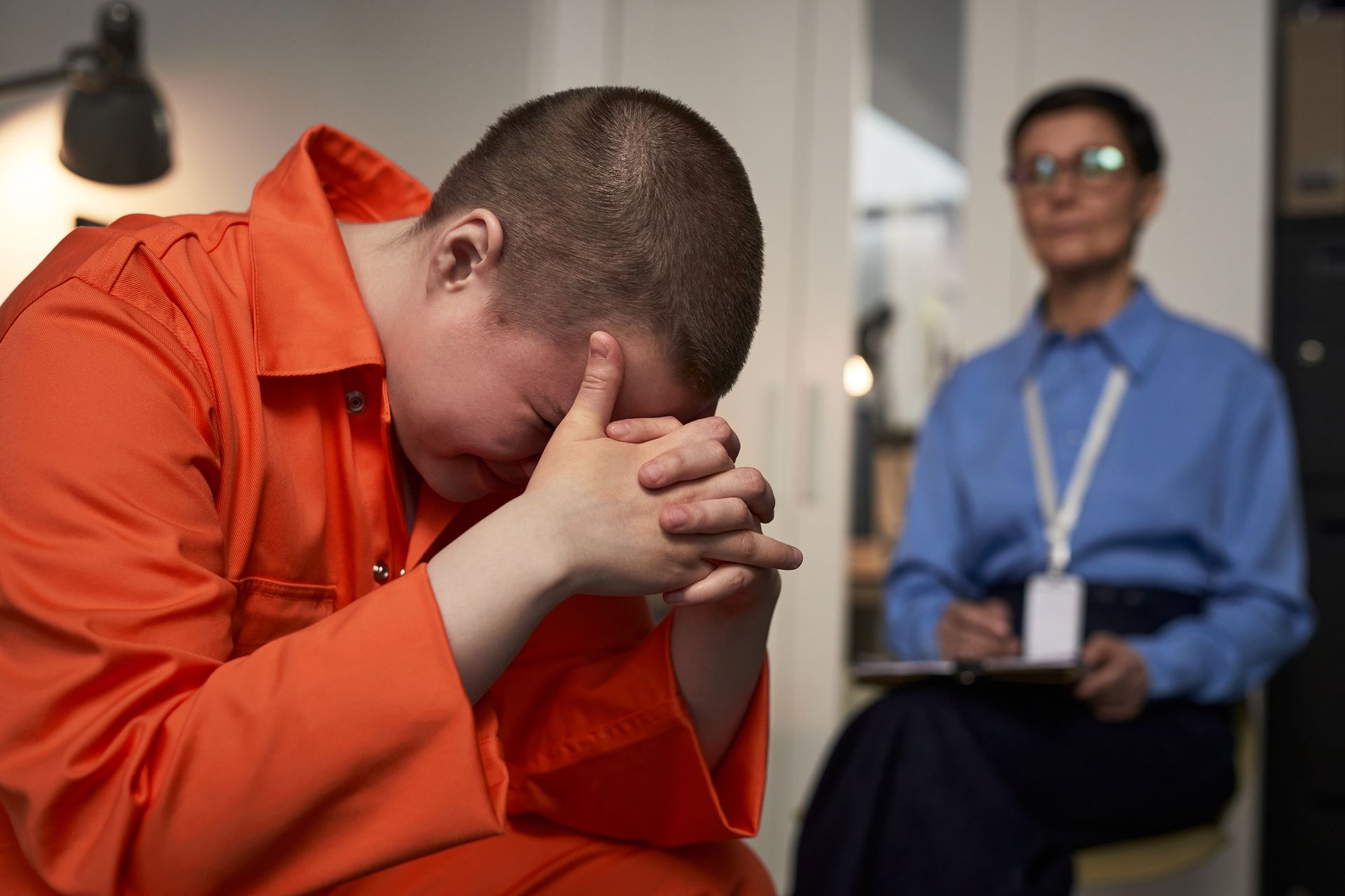Person in orange prison uniform with head in hands, crying, during therapy session. Therapist with notepad watches.
