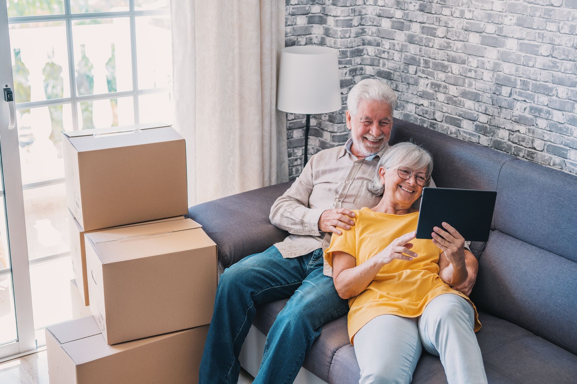 Elderly couple on a couch using a tablet, with moving boxes beside them in a bright living room.