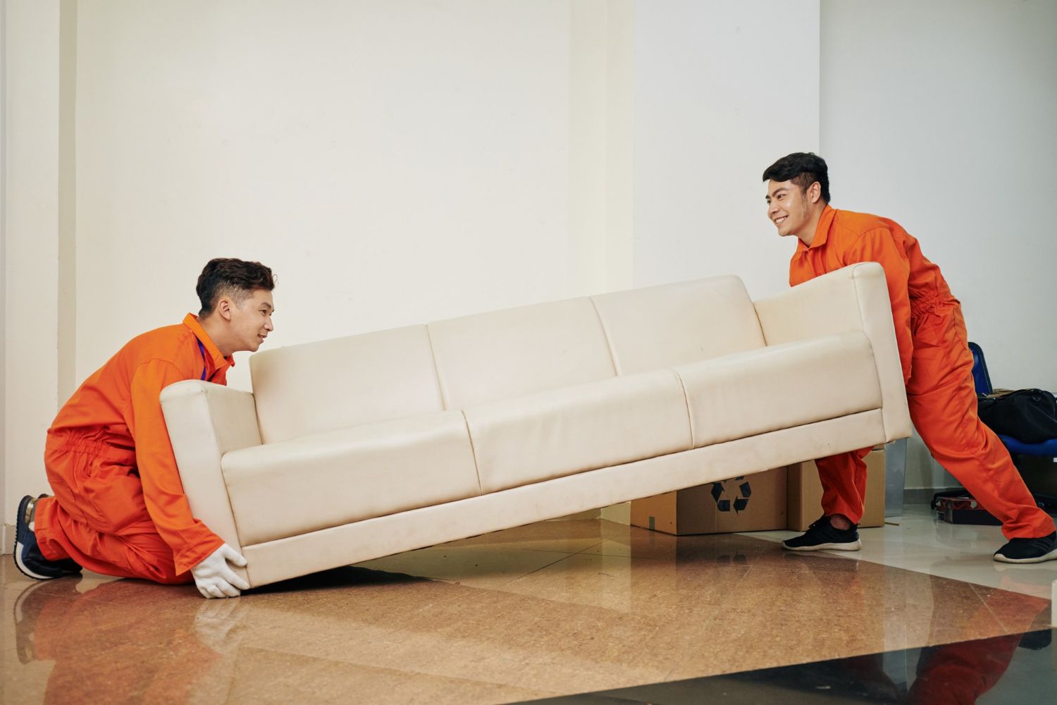 Two movers in orange coveralls lift a beige sofa in a brightly lit room.