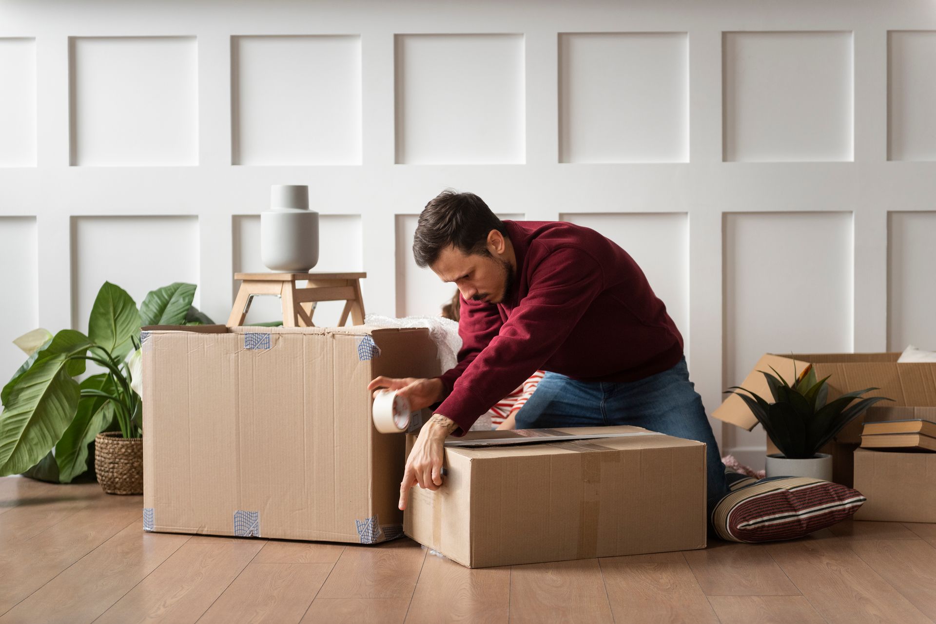 Man kneeling, sealing cardboard boxes with tape, preparing to move in a room with white paneling.