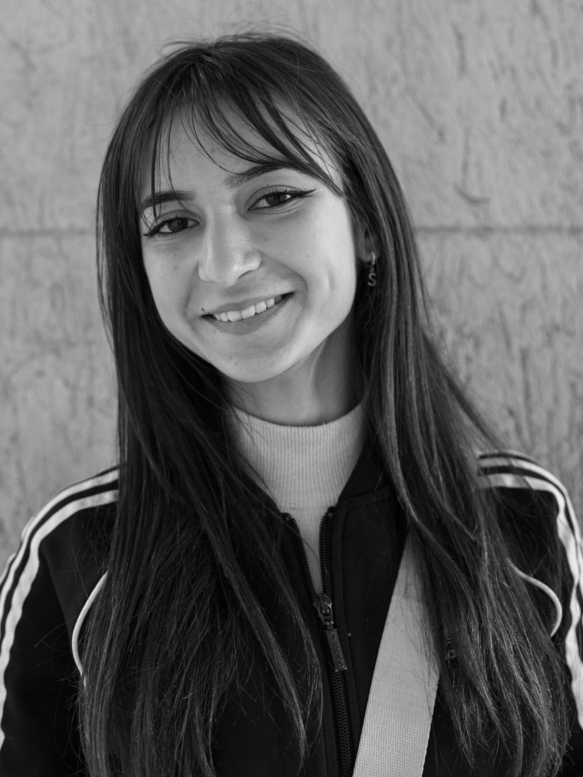 Woman with long dark hair, bangs, smiling, wearing a jacket with white stripes, and a white shirt.