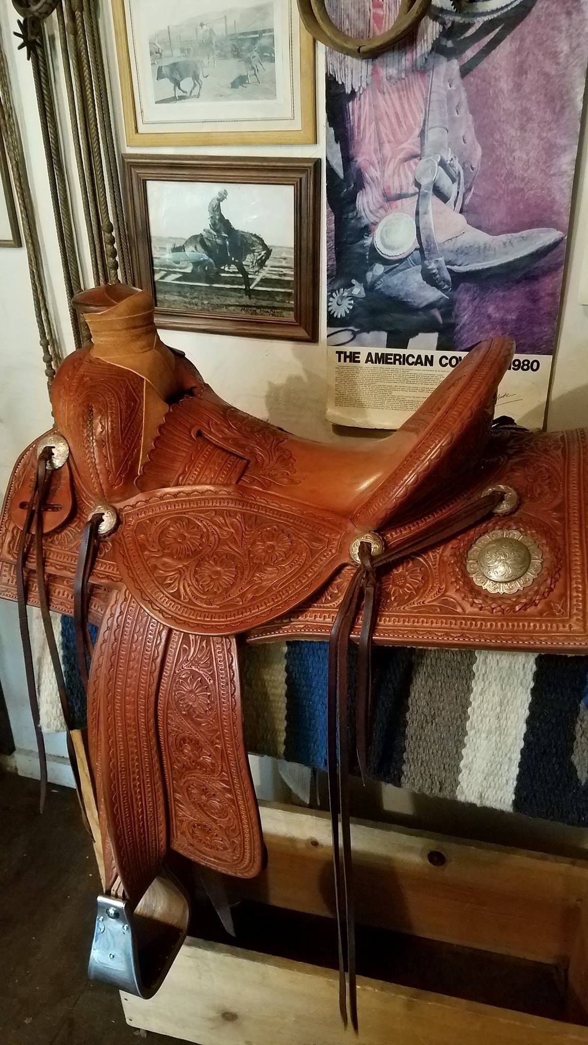 Brown western saddle displayed on a wooden table with framed wall art behind it.