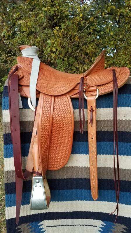 Brown leather saddle hanging on a striped blanket, with tack straps and a silver stirrup.