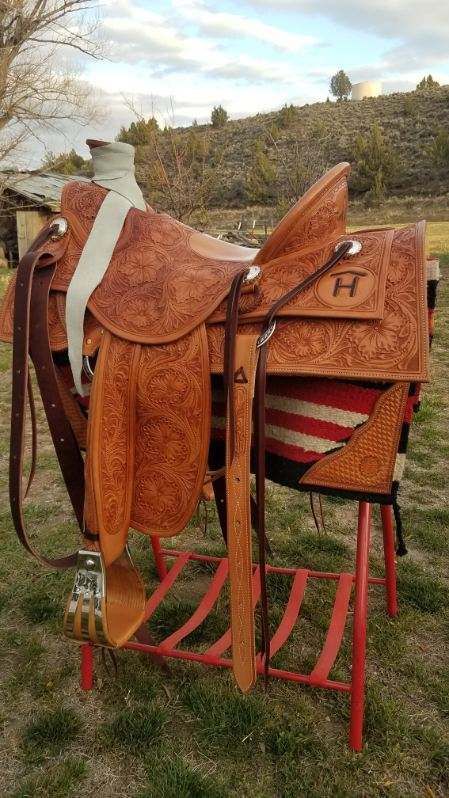 Brown leather saddle on red metal stand outdoors, with striped blanket and trees in the background