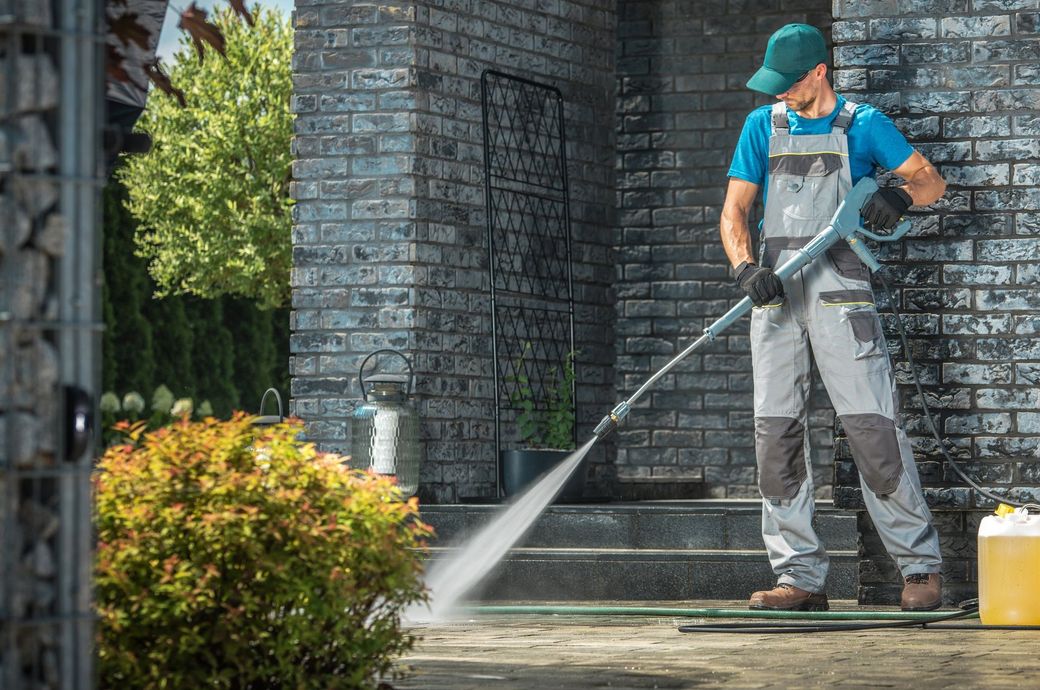 A person in work coveralls and a cap uses a power washer to clean a paved surface in front of a brick house.