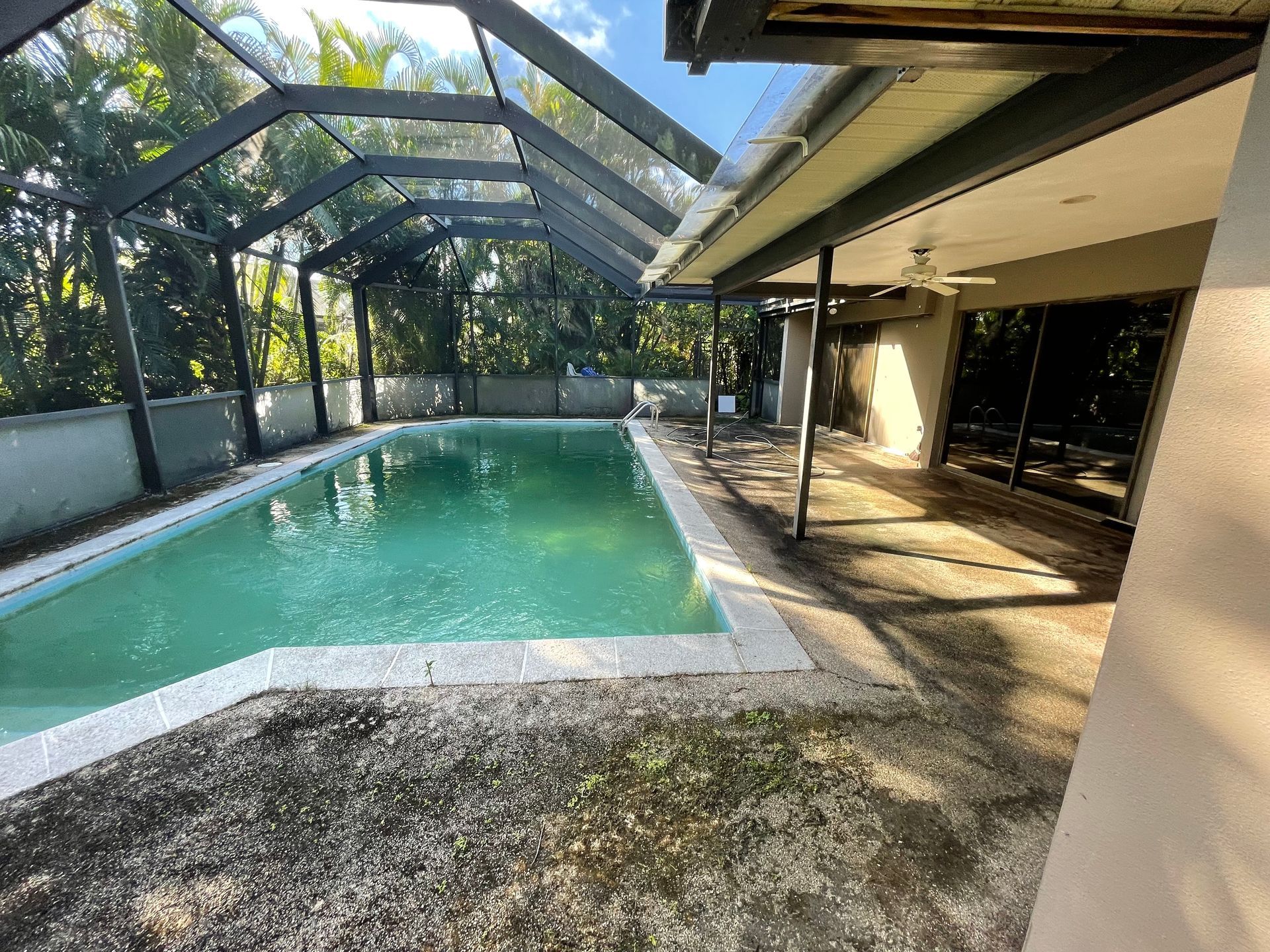 A rectangular swimming pool with murky water inside a screened-in lanai attached to a home.