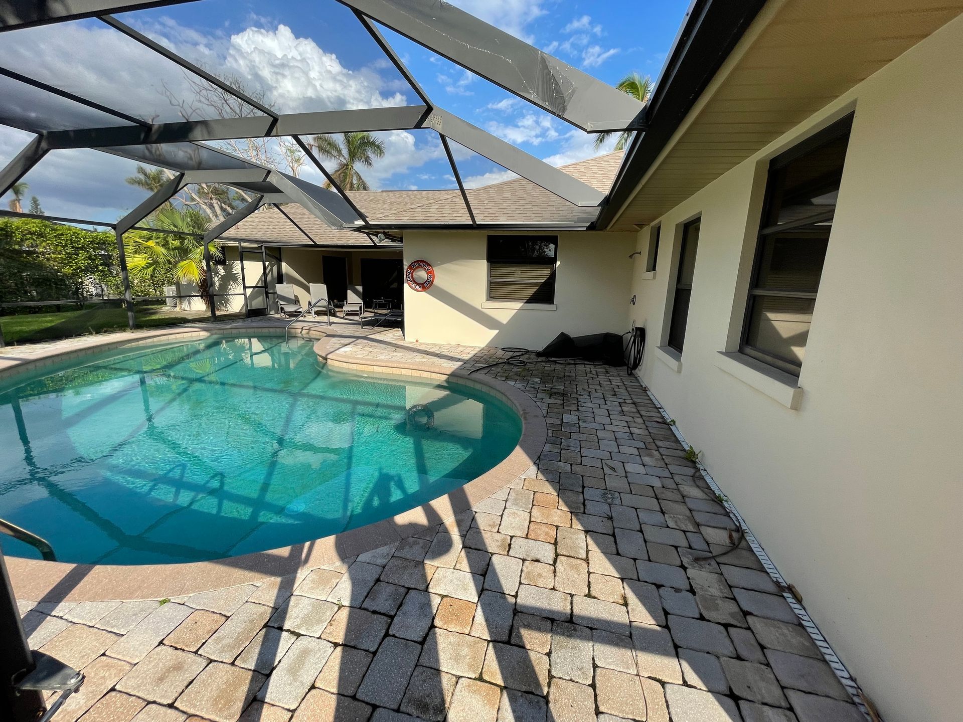 A swimming pool with a screened enclosure, stone patio, and a house exterior on a sunny day.