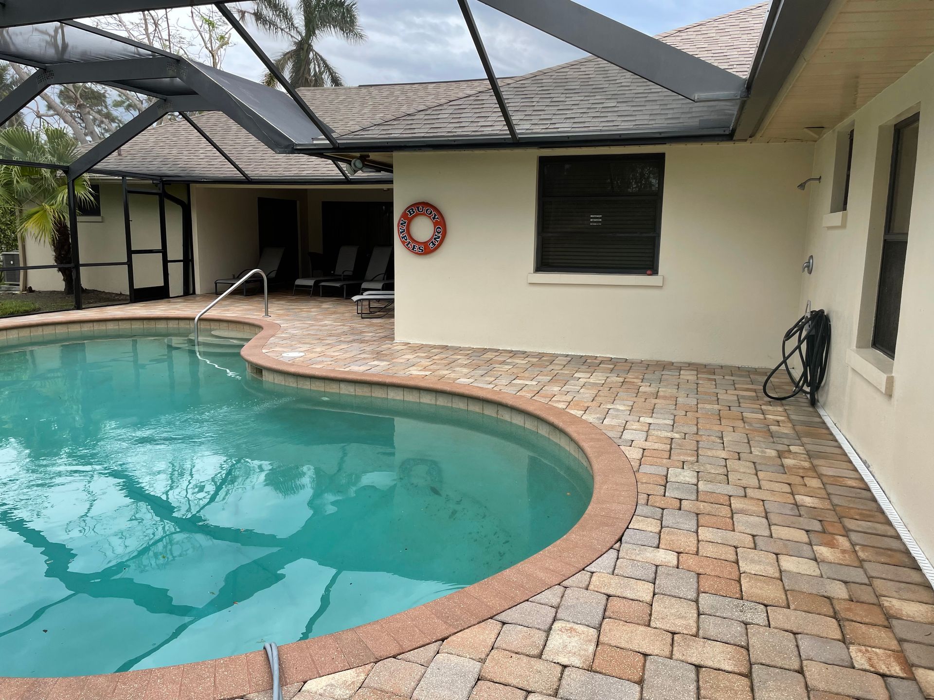 A backyard swimming pool area with beige stone pavers, a screened-in enclosure, and a life preserver on the wall.