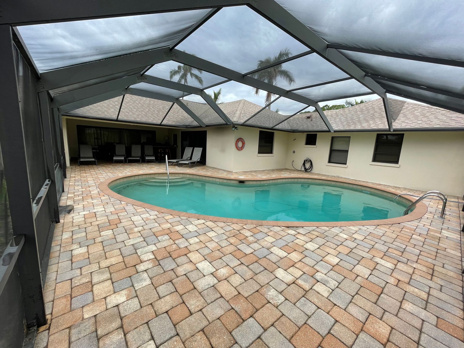 A backyard pool with a kidney-shaped basin, surrounded by tan pavers and protected by a full-coverage screen enclosure.