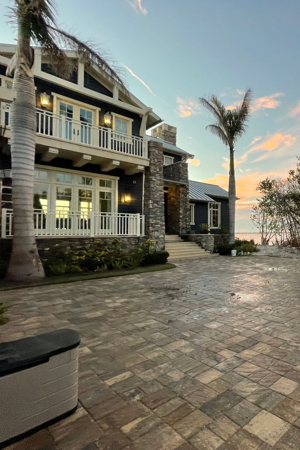 A two-story coastal home with stone siding and a paved driveway at sunset, featuring palm trees and a balcony.