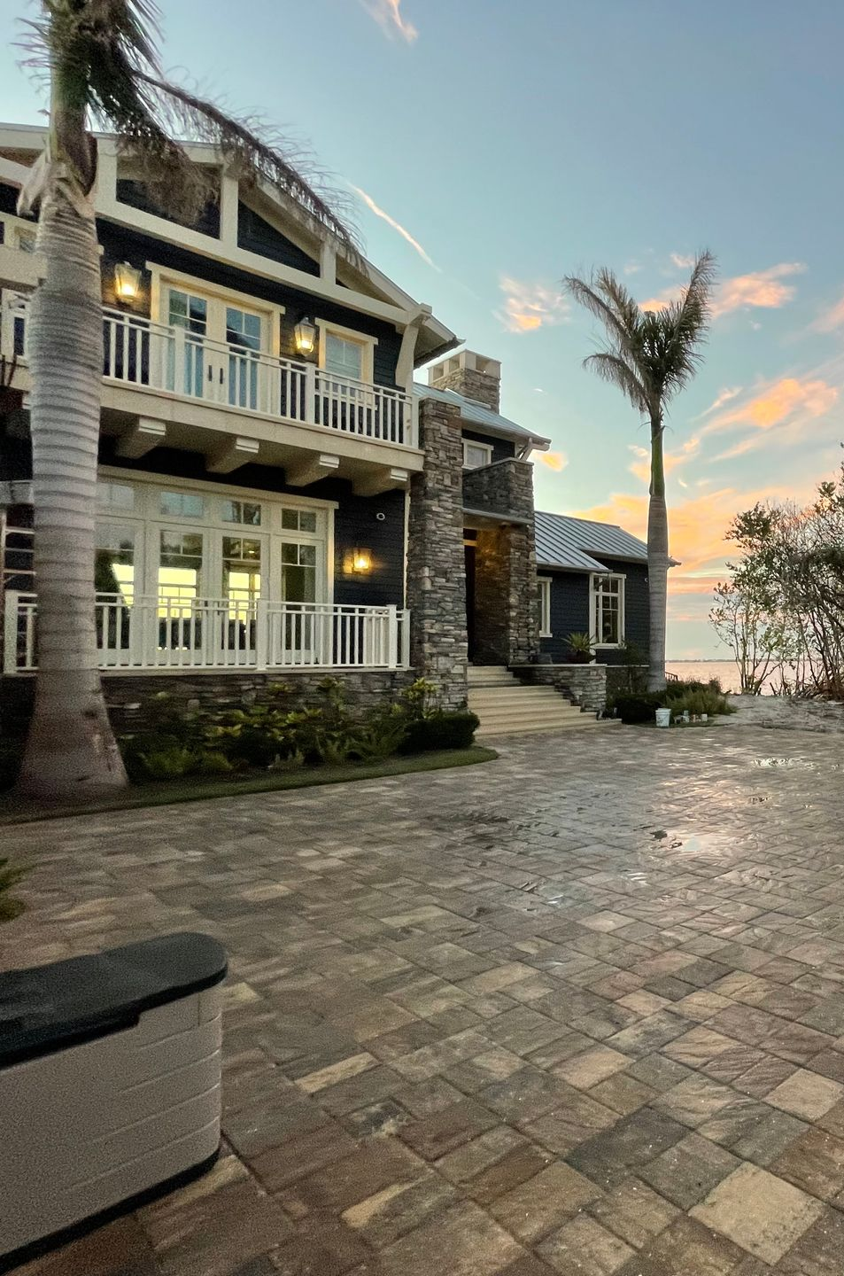 A two-story dark-sided coastal home with a stone chimney and stone patio at dusk, framed by two tall palm trees.
