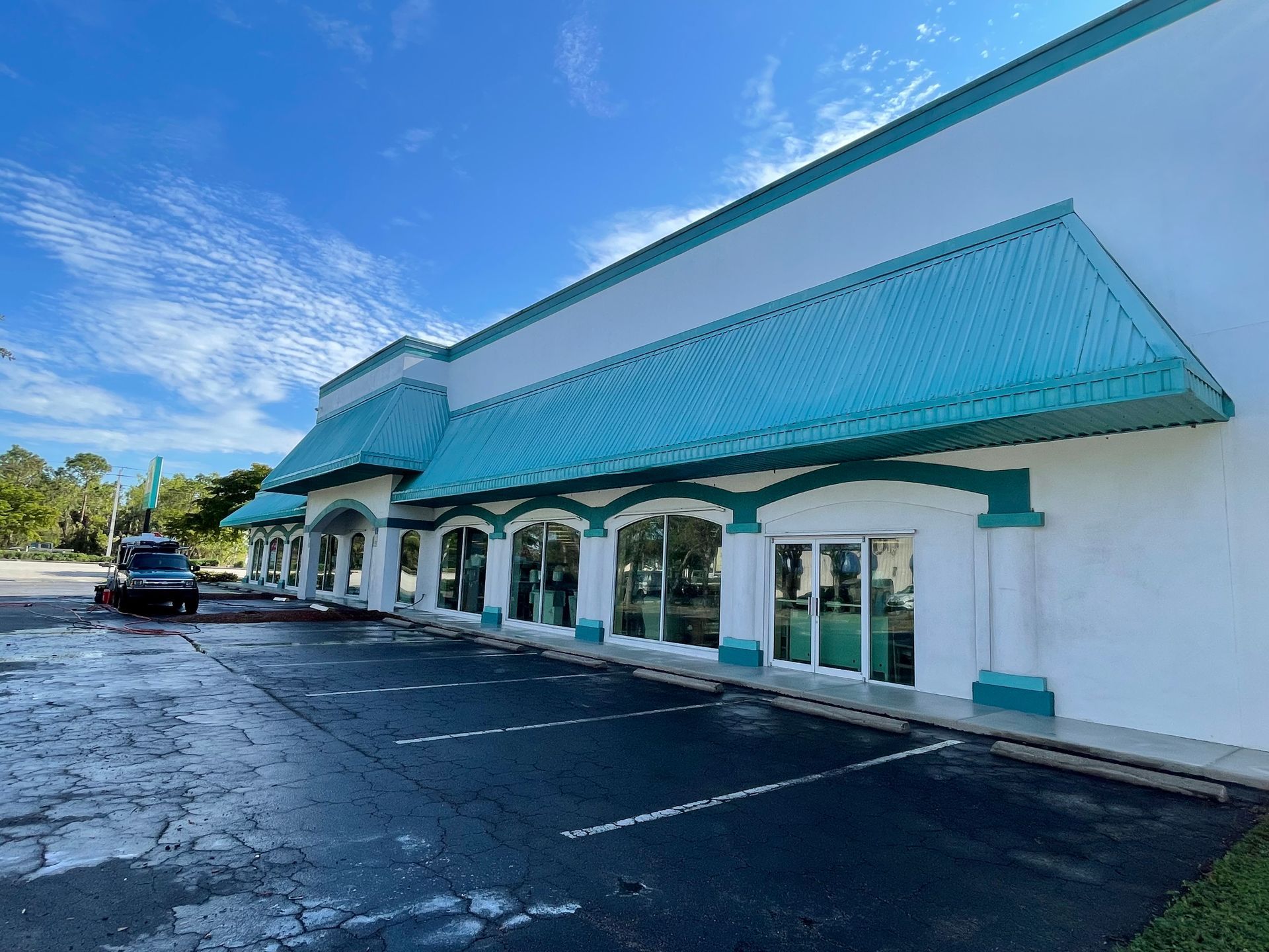A white commercial building with a long teal awning and pillars, seen from the corner of a mostly empty parking lot.
