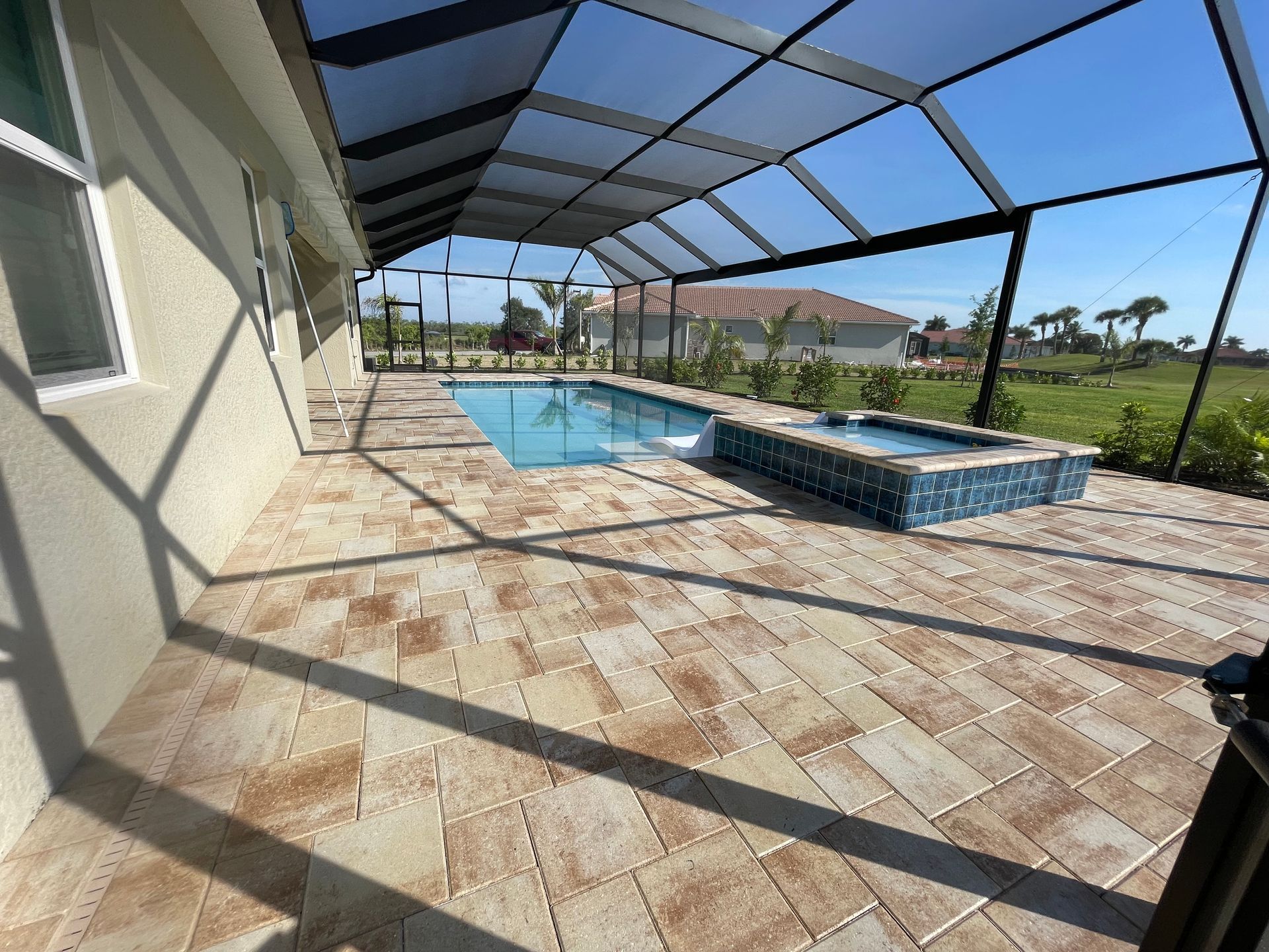 A sunny outdoor pool and raised hot tub enclosed by a screen, featuring light-colored stone pavers on the patio deck.