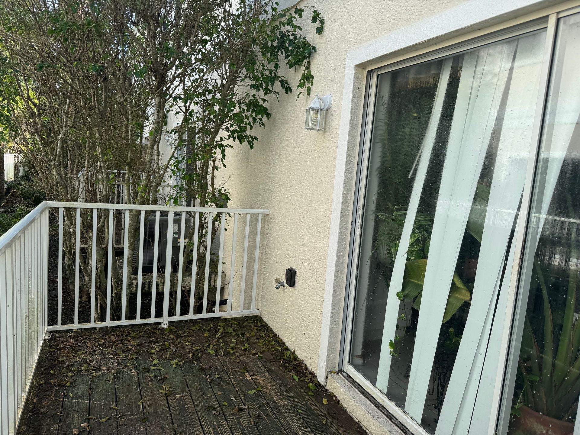 A small balcony with wood-plank flooring, a white metal railing, an AC unit behind a tree, and a sliding glass door.