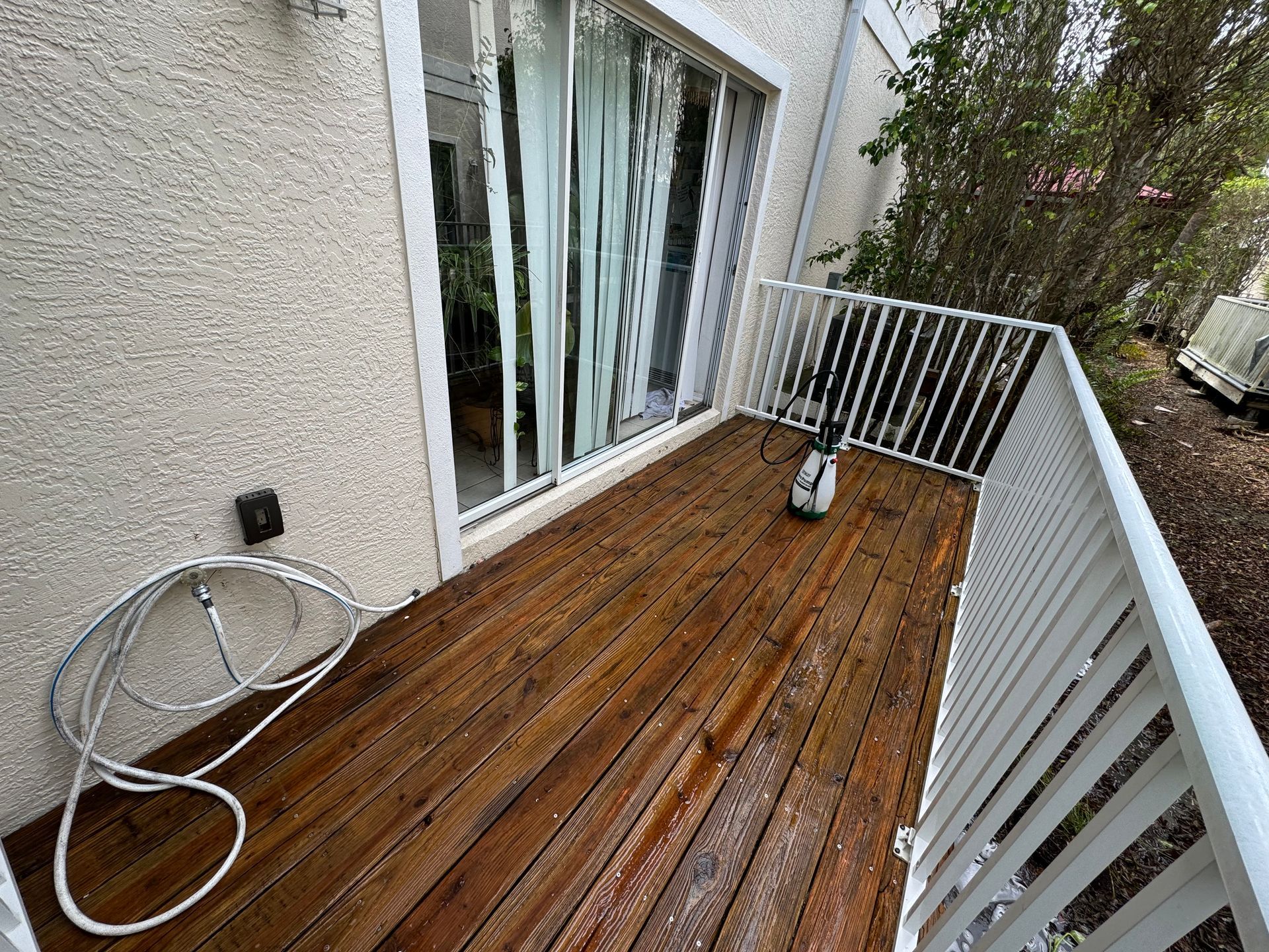 A wooden deck with a white railing and a sliding glass door, featuring a coiled hose on the left and a spray nozzle.