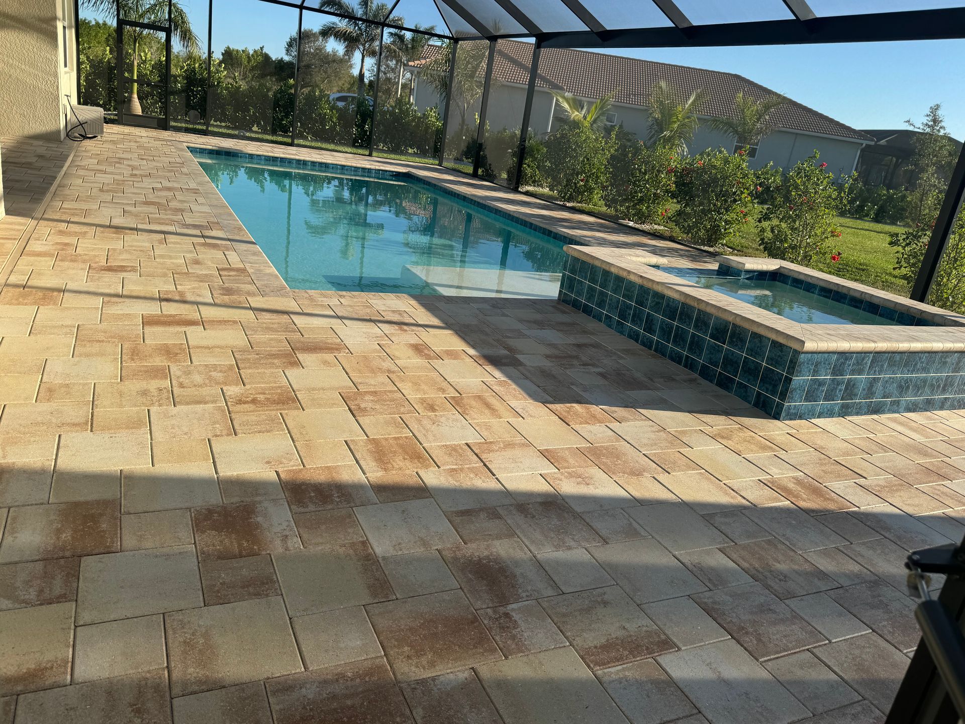 A residential swimming pool and attached spa on a patio with tan stone pavers under a screen enclosure.