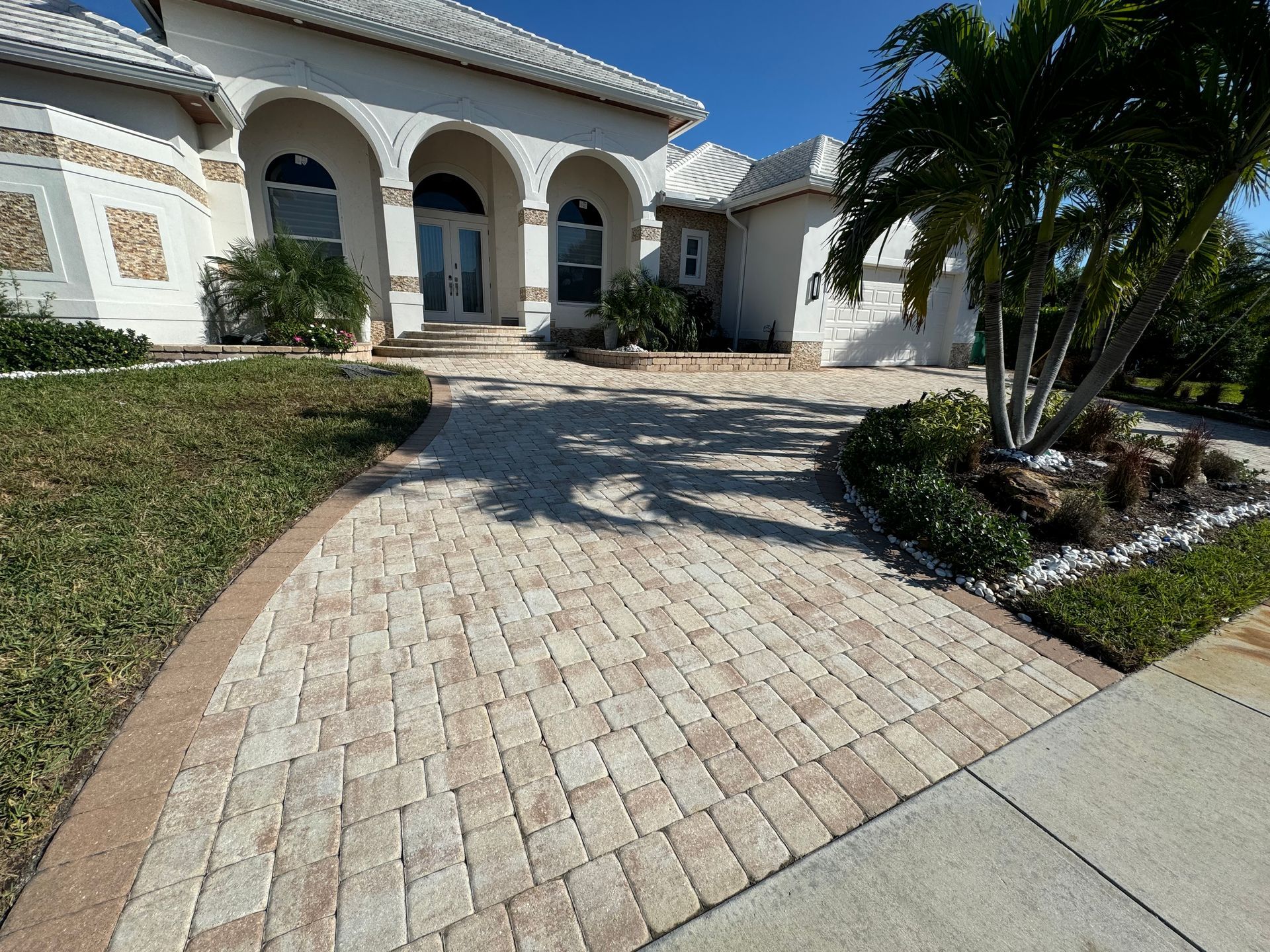 A light-colored brick paver driveway leads to the arched entryway of a white stucco house on a sunny day.