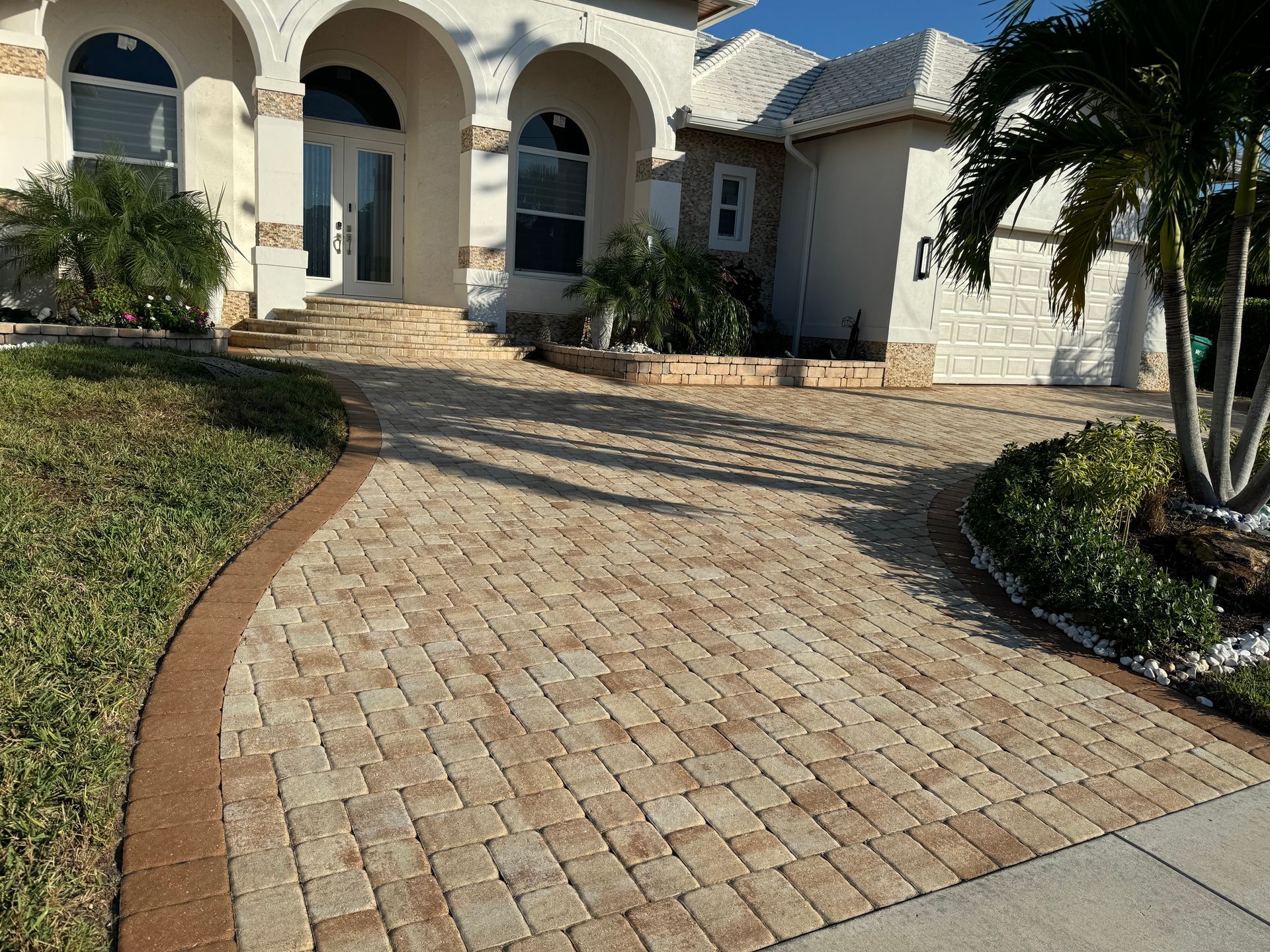A house with a beige brick paved driveway leading to an arched entryway, surrounded by grass and small palm trees.