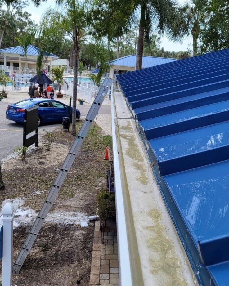 A ladder leans against a building with a blue metal roof and a long, debris-filled gutter, outdoors near a parking lot.