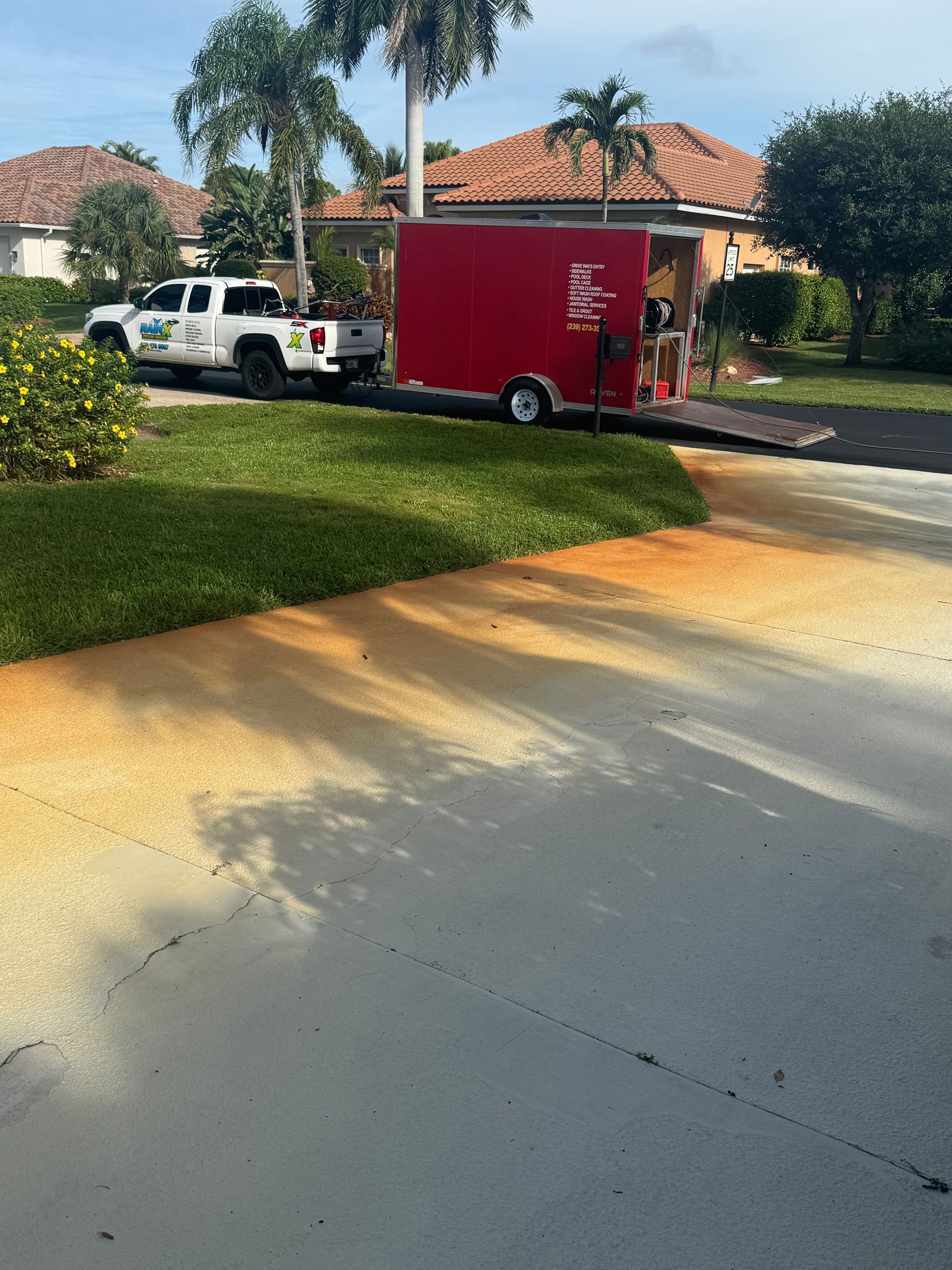 A white pickup truck with a red cargo trailer parked on a driveway with prominent rust-colored stains on the concrete.