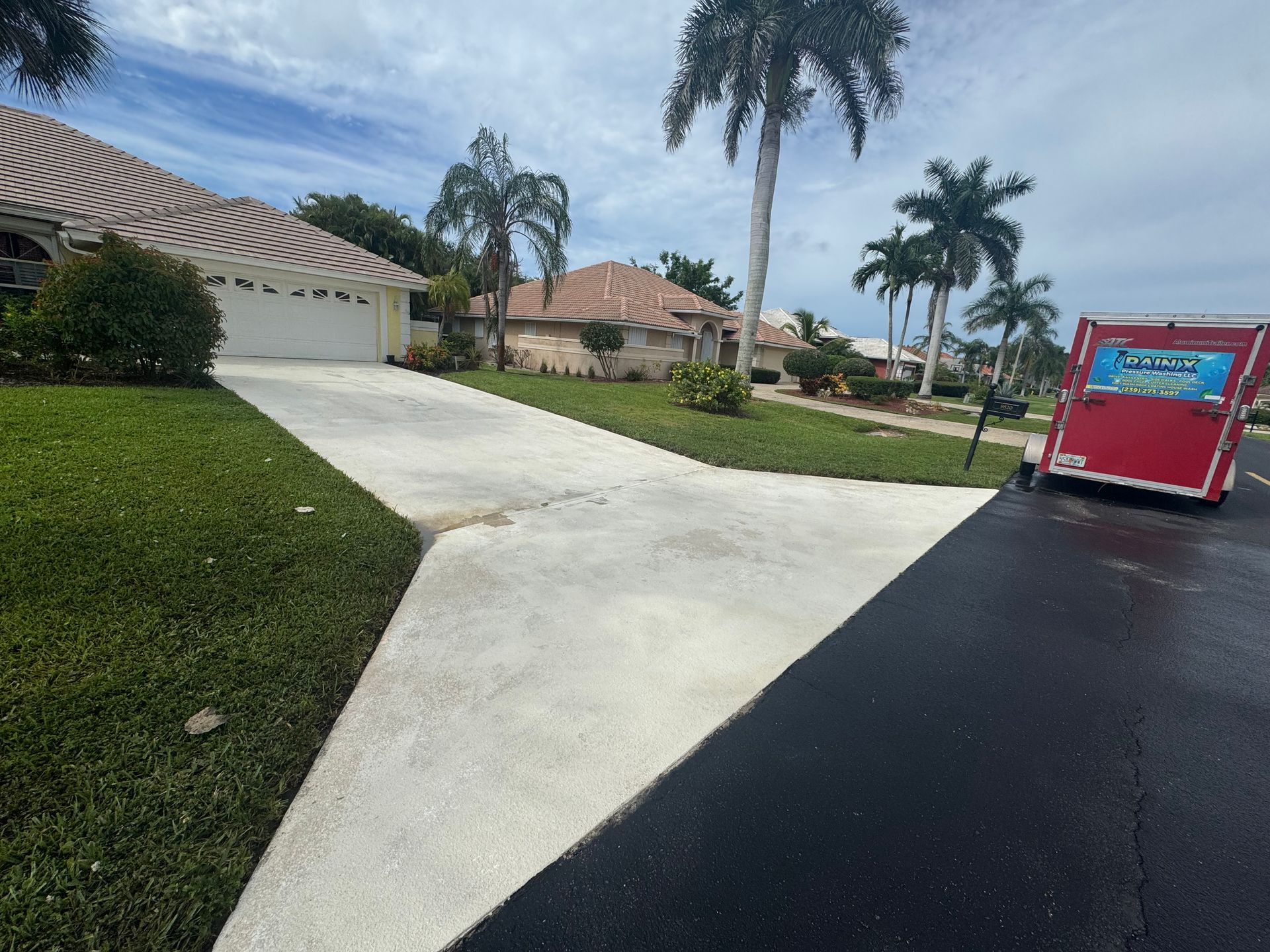 A concrete driveway leading to a suburban house with a red trailer parked on the adjacent asphalt street.