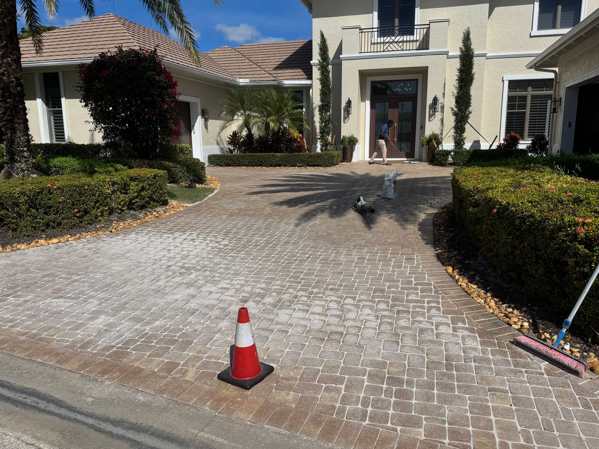 A paved driveway leading to a large house, featuring a construction cone in the foreground and a broom to the right.