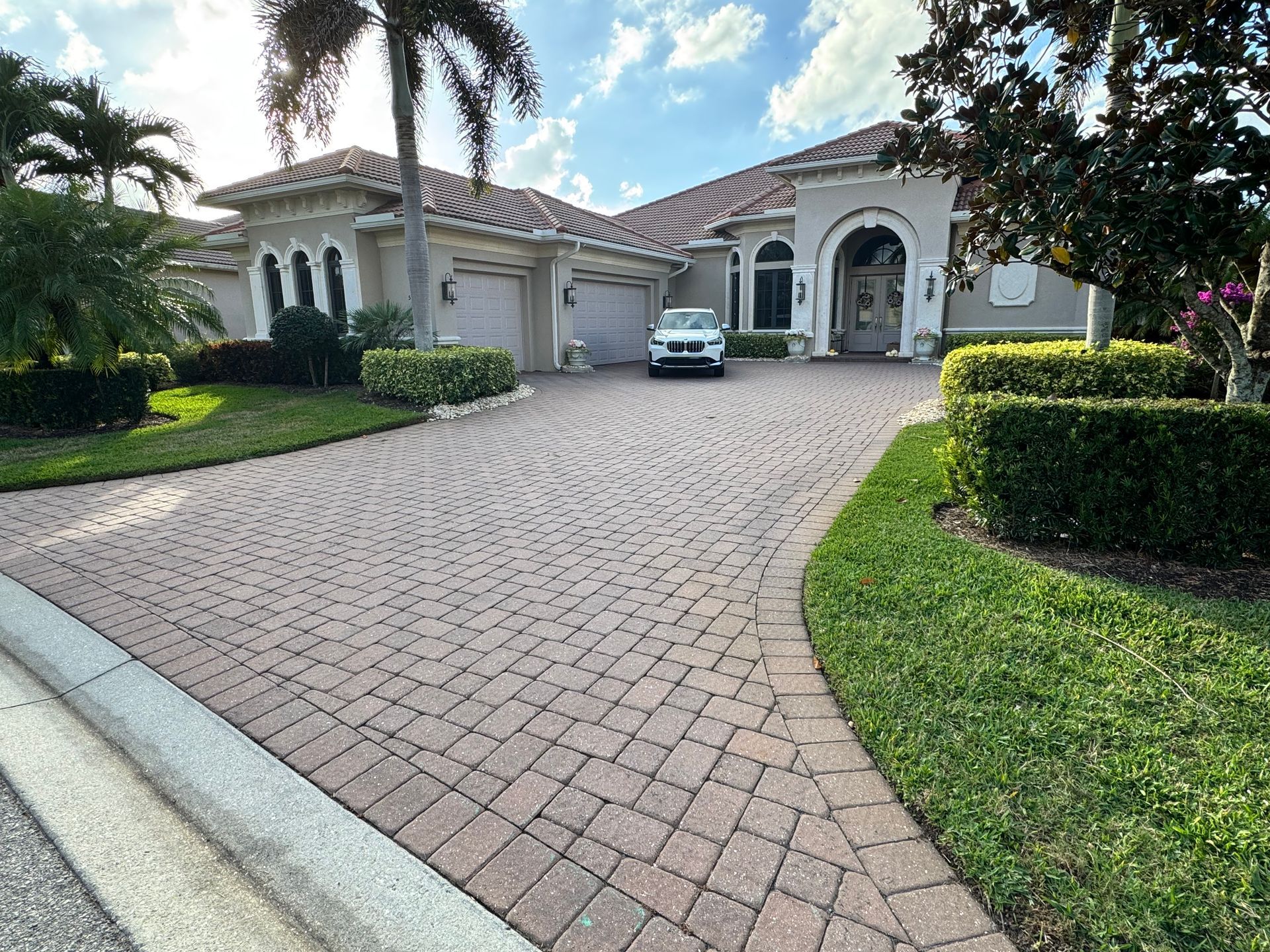 A luxury house with a large paved driveway, manicured landscaping, and a car parked in front of the arched entryway.