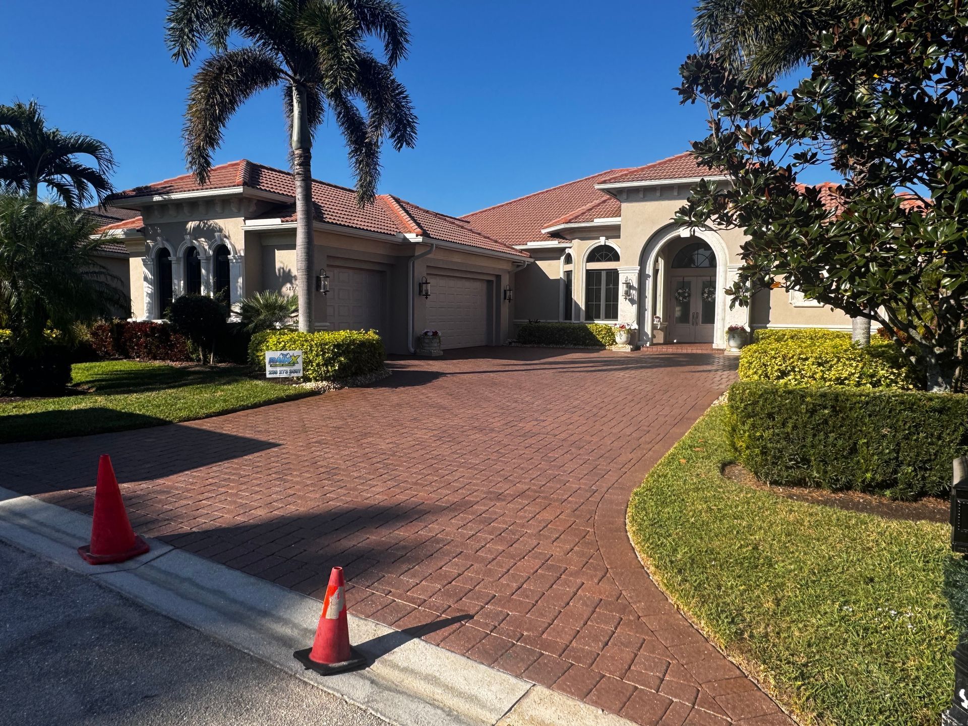 A beige single-story house with a red-tiled roof and a brick driveway, featuring two orange cones near the street.